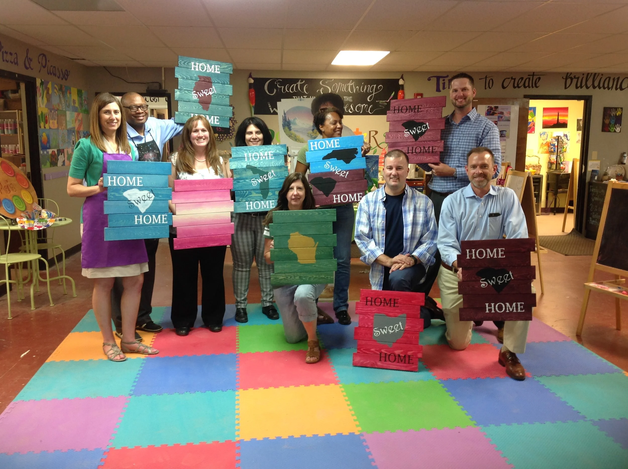 A group of ten people posing indoors with colorful wooden signs that read 'Home' and 'Sweet'. Some signs also have the outline of Illinois or the state of Wisconsin painted on them. The background features various art displays and a positive quote on