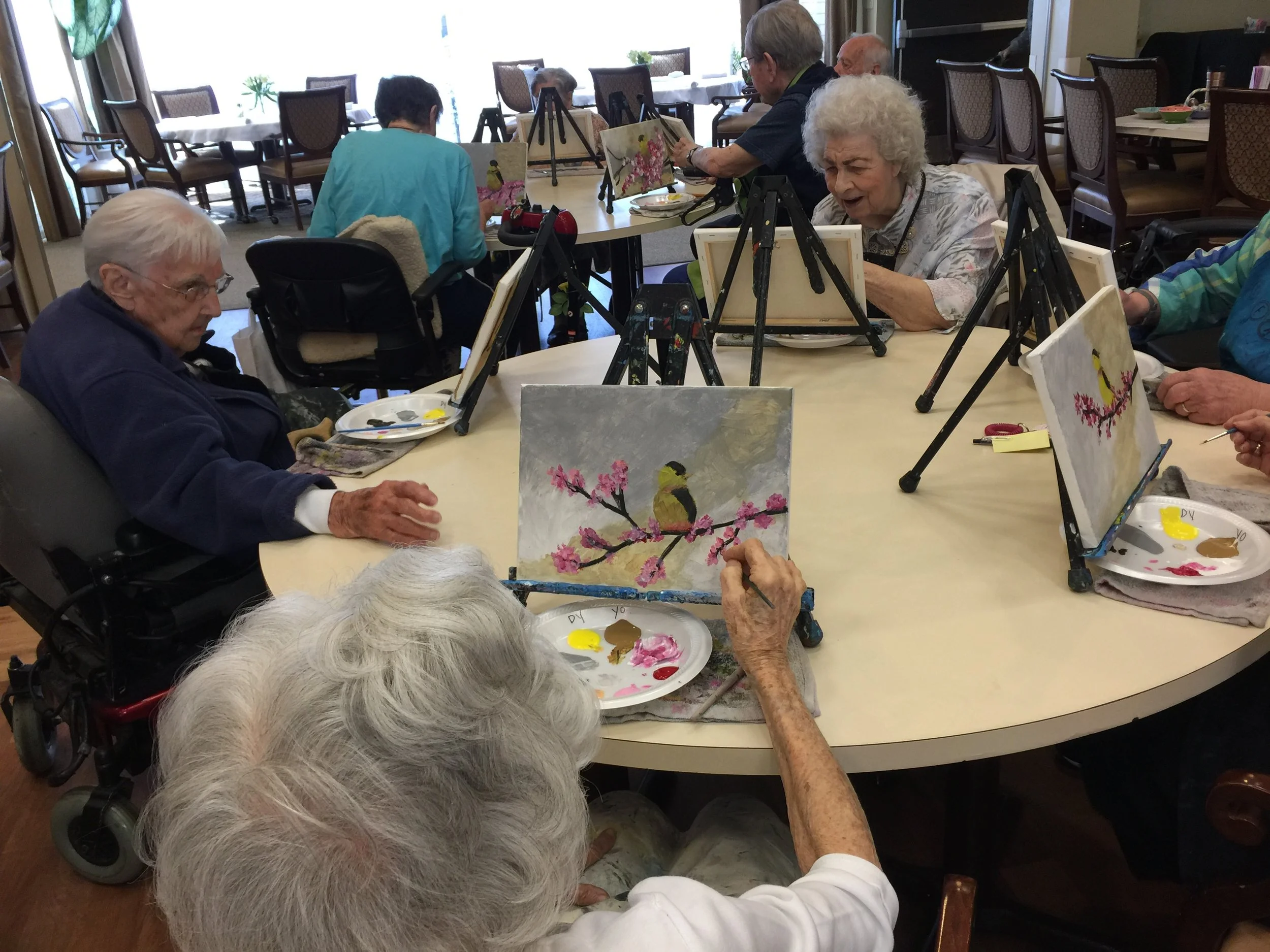 Elderly people participating in a group painting activity at a table, creating artworks of a bird on a cherry blossom branch.