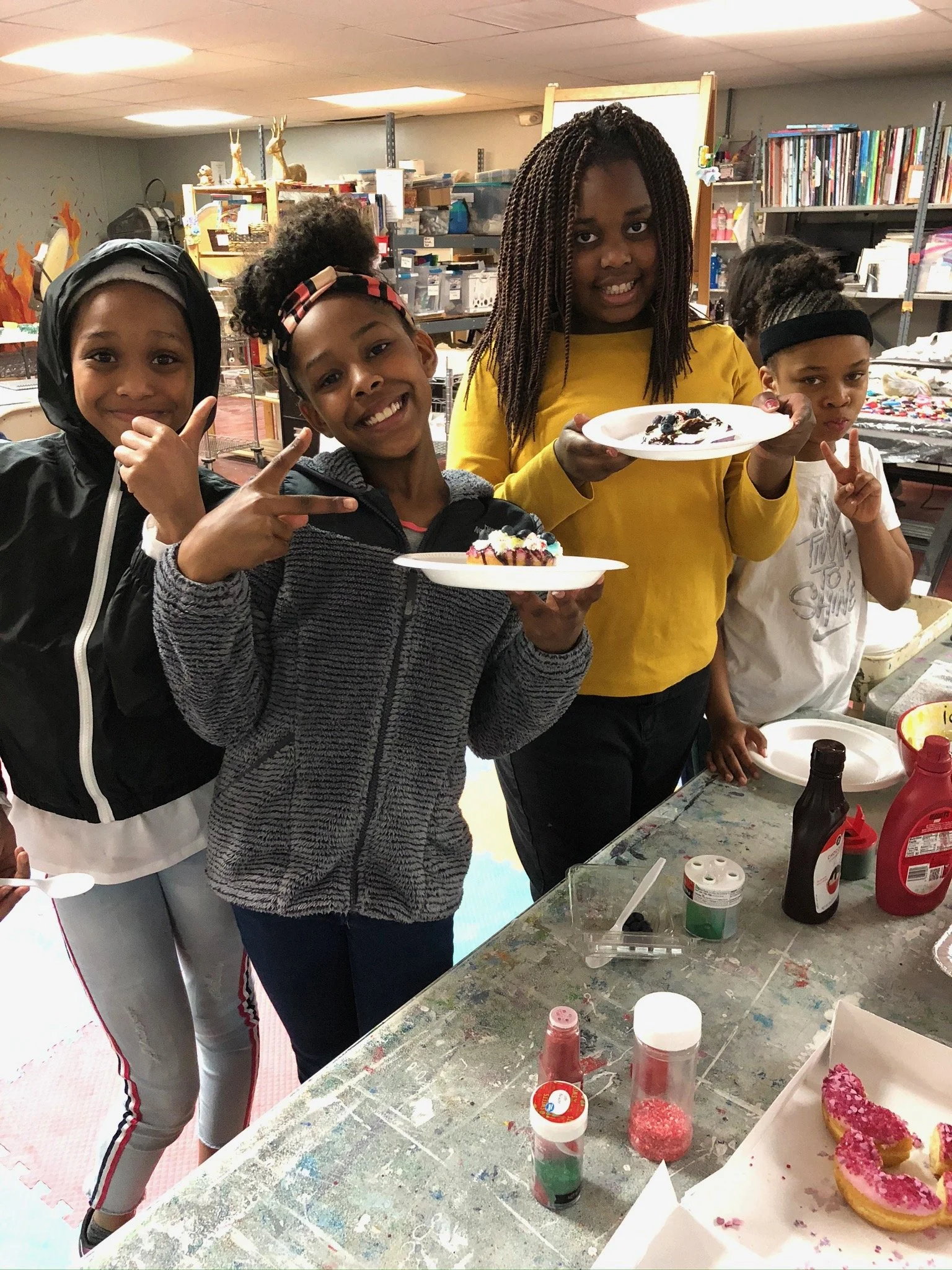 Four young girls celebrating with decorated slices of cake in a colorful art classroom or craft room.