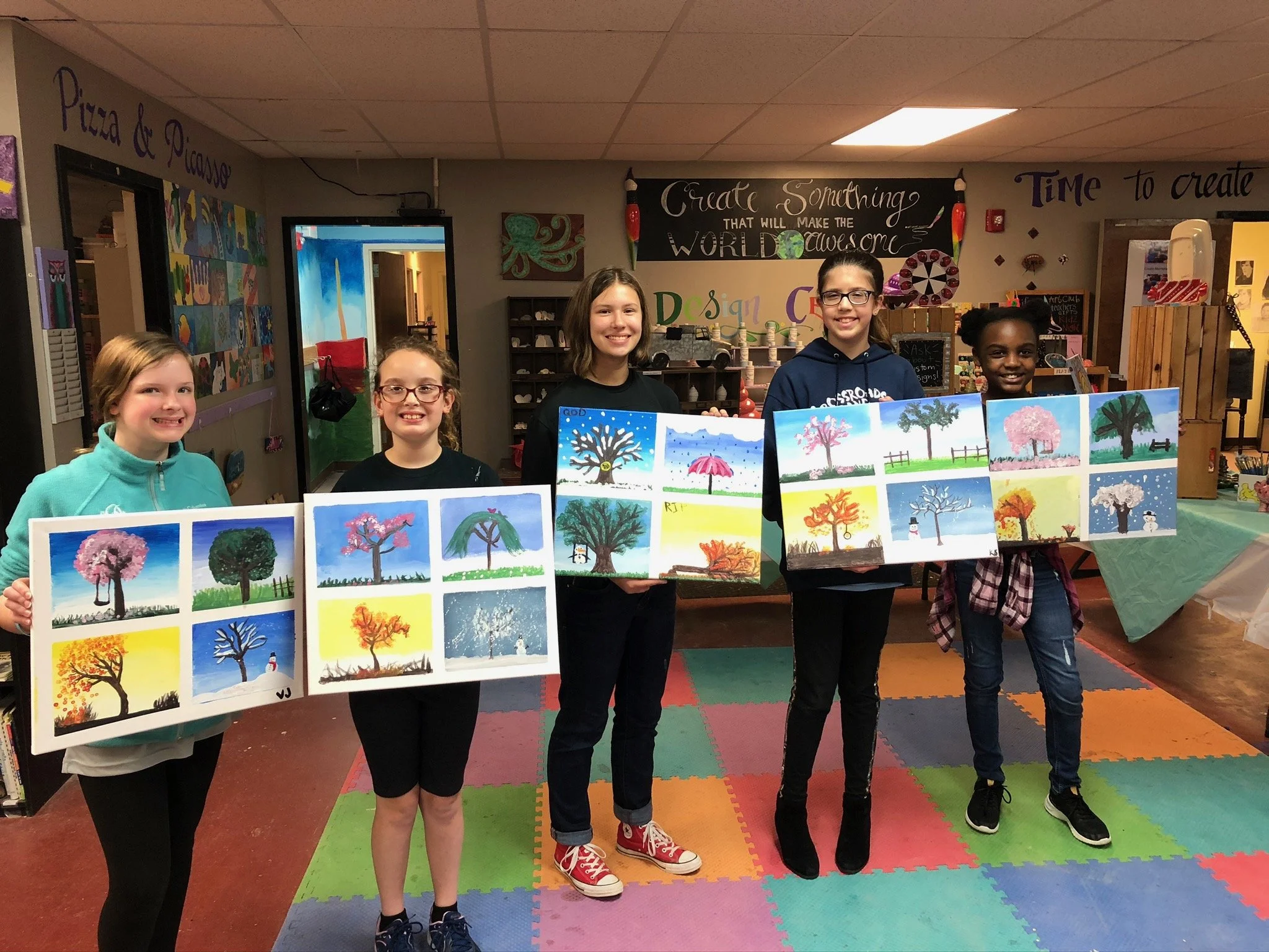 Five girls standing in a classroom holding colorful paintings of trees and seasons. The classroom has colorful mats on the floor and artwork and signs on the walls.