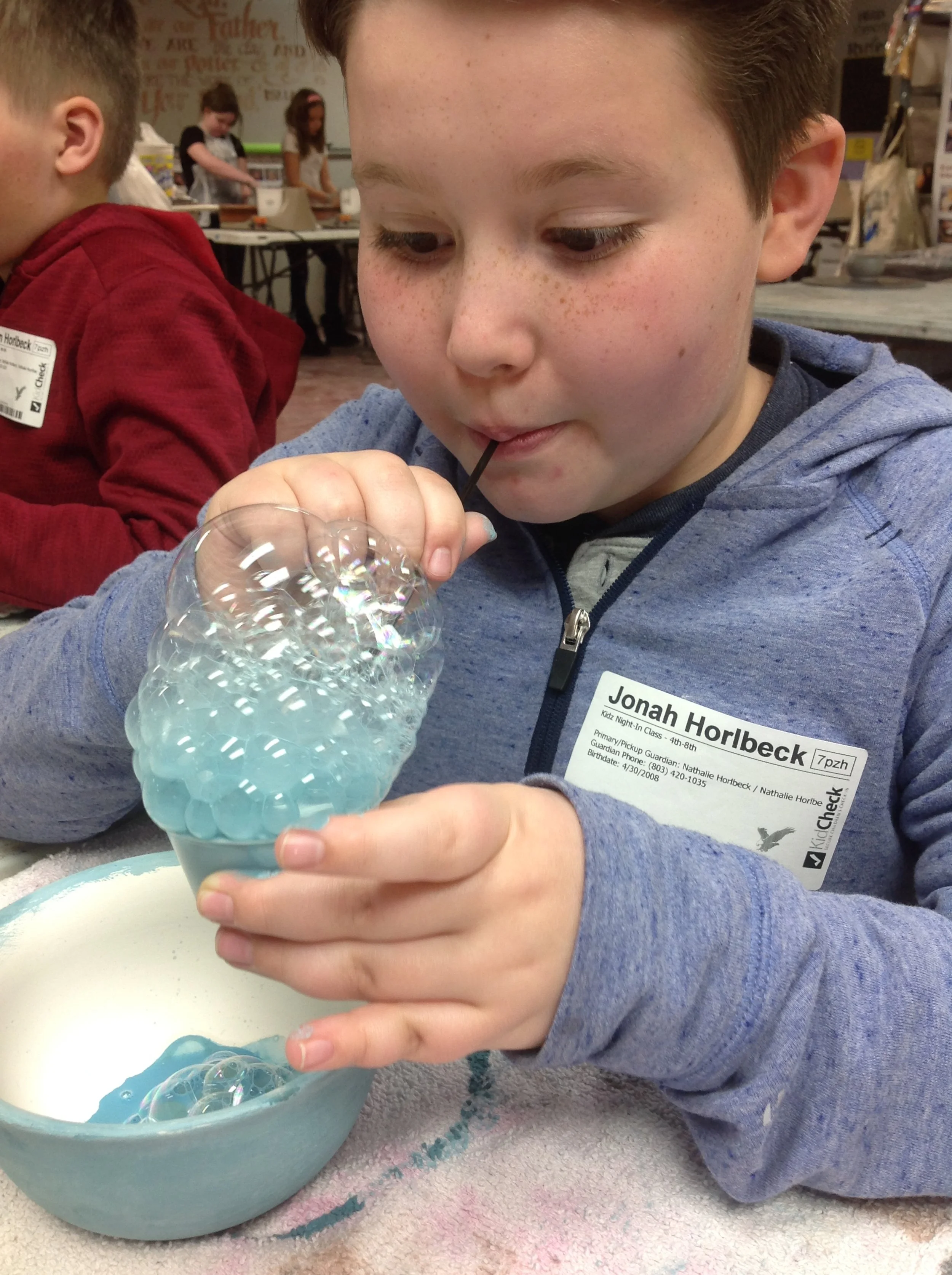 Young boy blowing bubbles in a classroom or activity center, with other children and educational decor in the background.