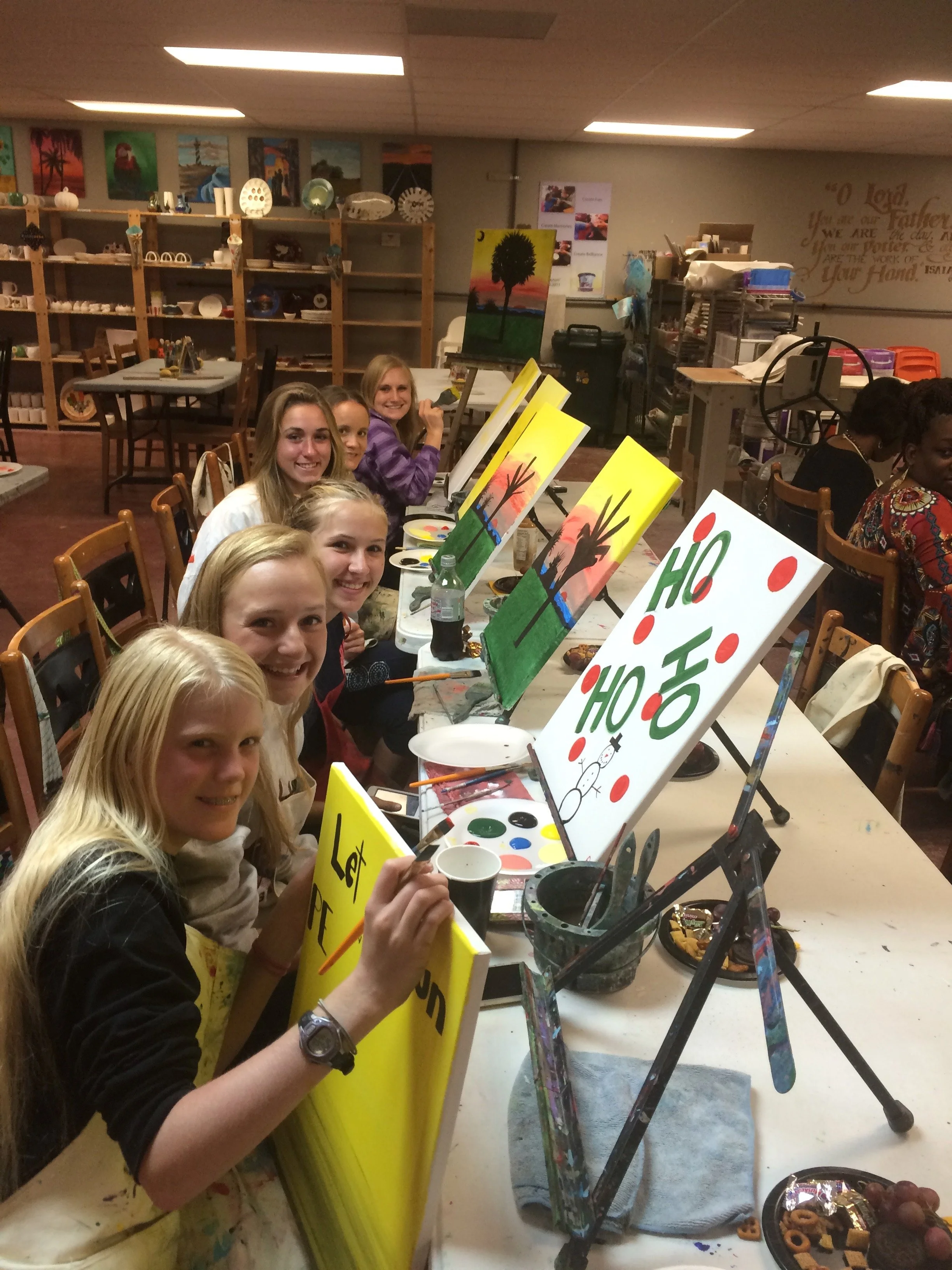 Six young women sitting at a table painting colorful holiday-themed canvases with reindeer, Christmas trees, and 