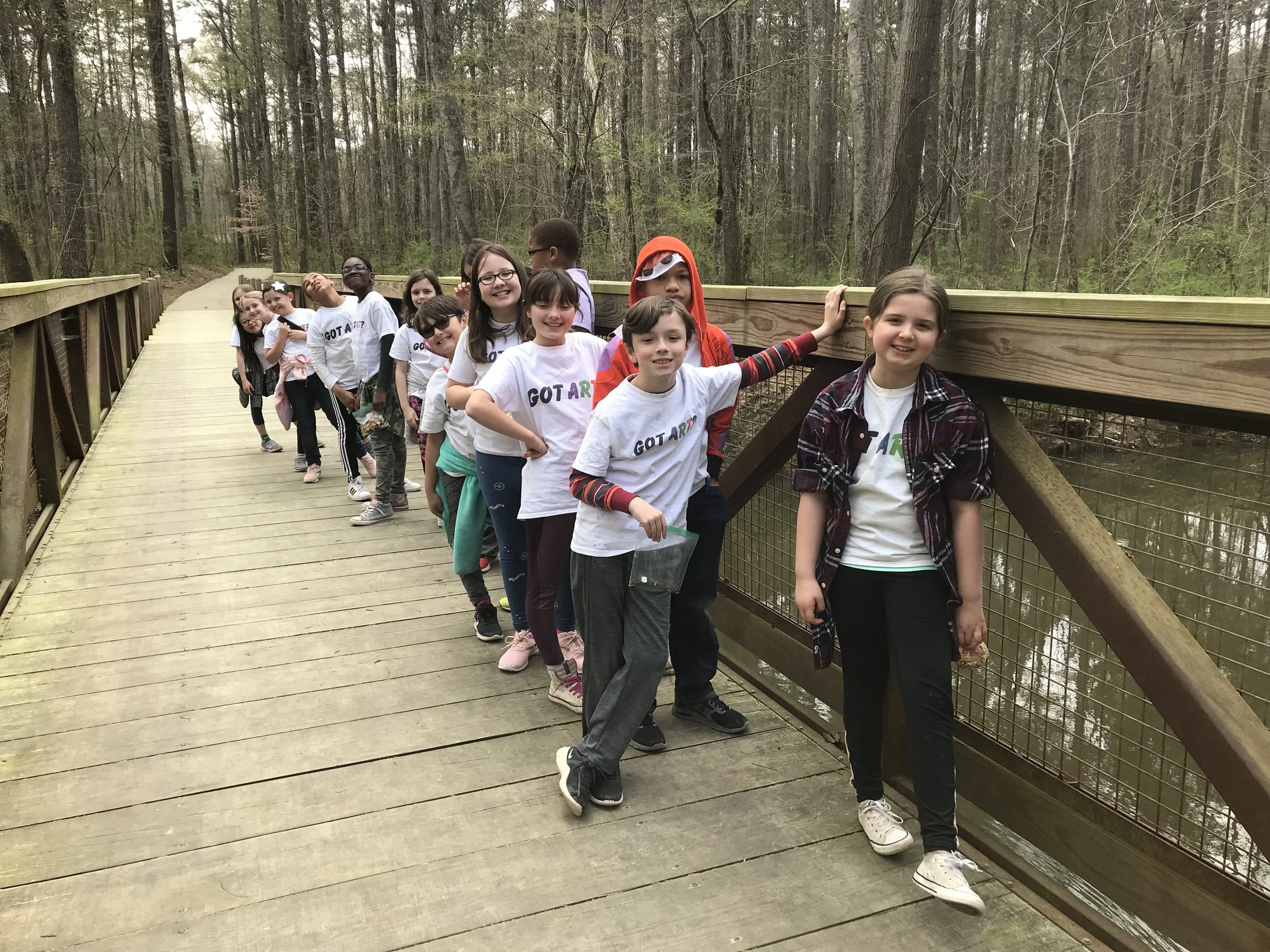 A group of children standing in a line on a wooden bridge in a forest, smiling and wearing white t-shirts with 'GOT AR' written on them.