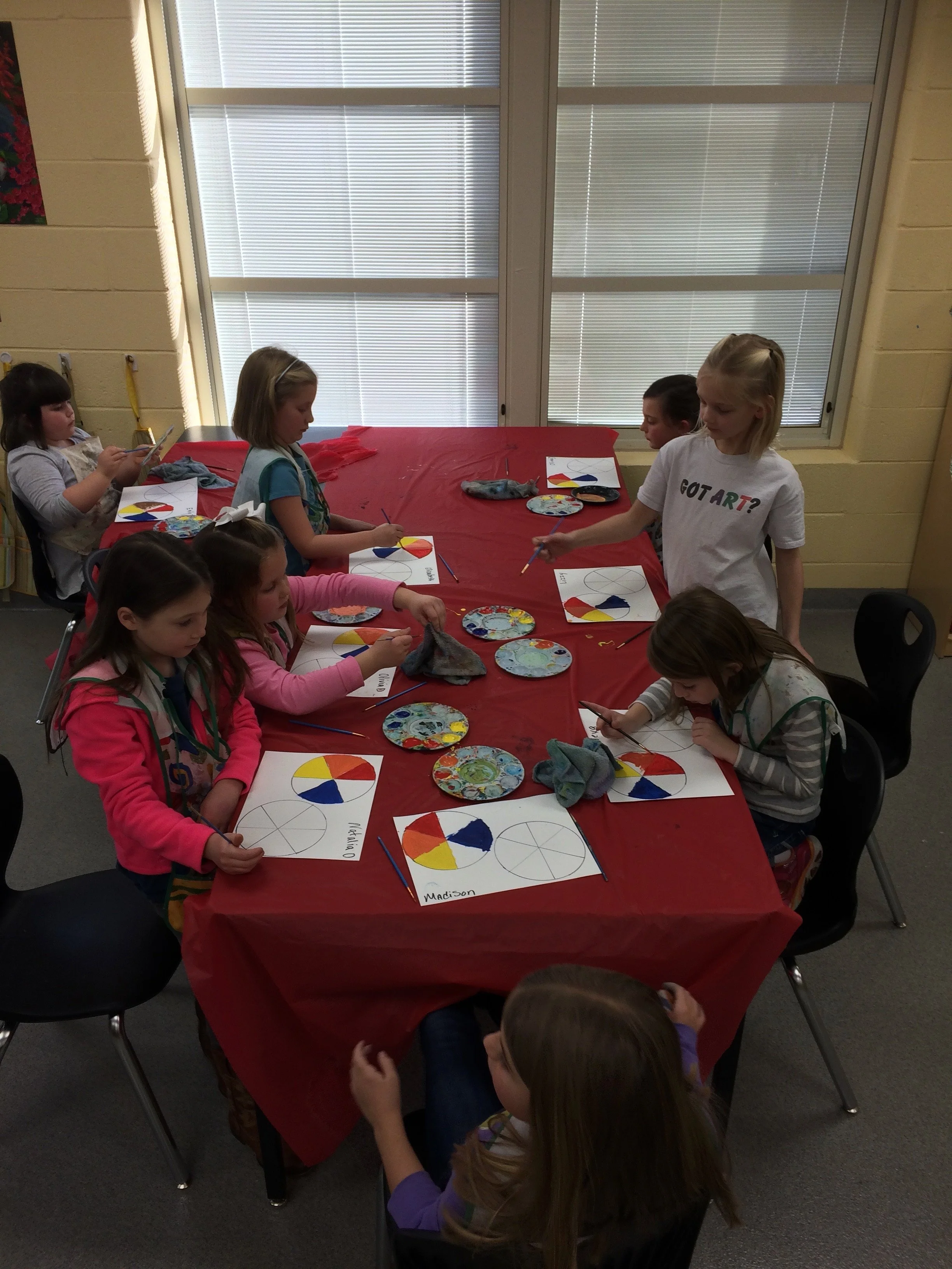 Children participating in a pie chart art project around a red table, each with a paper, brushes, and colorful plates of paint, in a classroom with large windows.