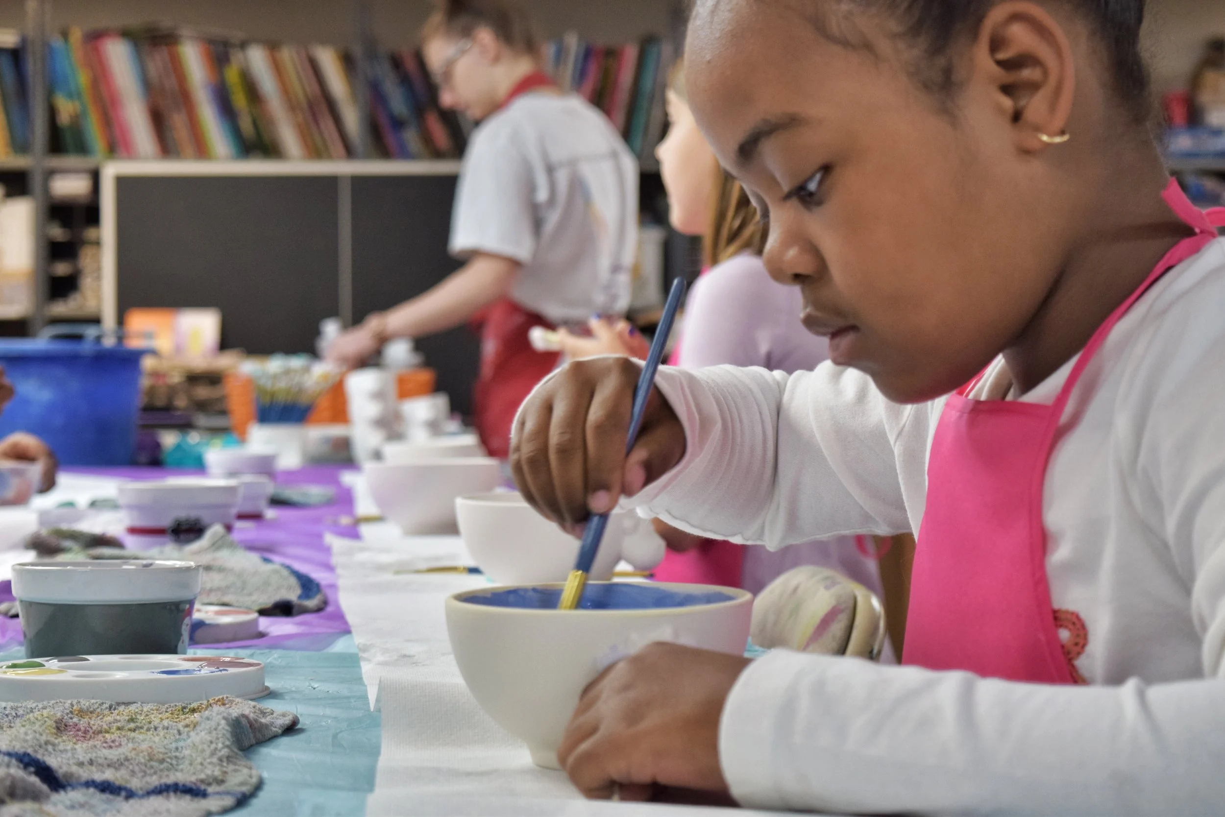 Children participating in a pottery painting activity, wearing aprons, in a classroom or art studio with shelves of books and art supplies in the background.