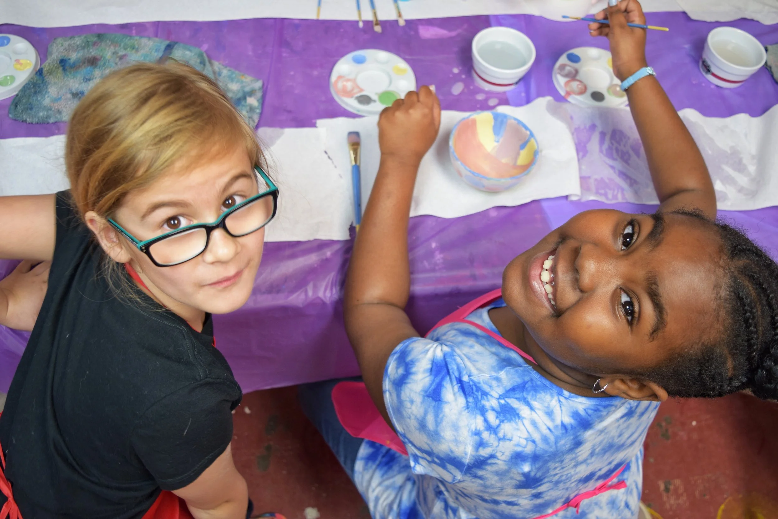 Two young girls smiling while painting with craft supplies on a purple tablecloth.