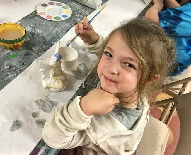 A young girl with light brown hair, smiling and resting her chin on her hand, sits at a table with ceramic painting supplies, including a white ceramic figure, a cup, a paint palette, and brushes.