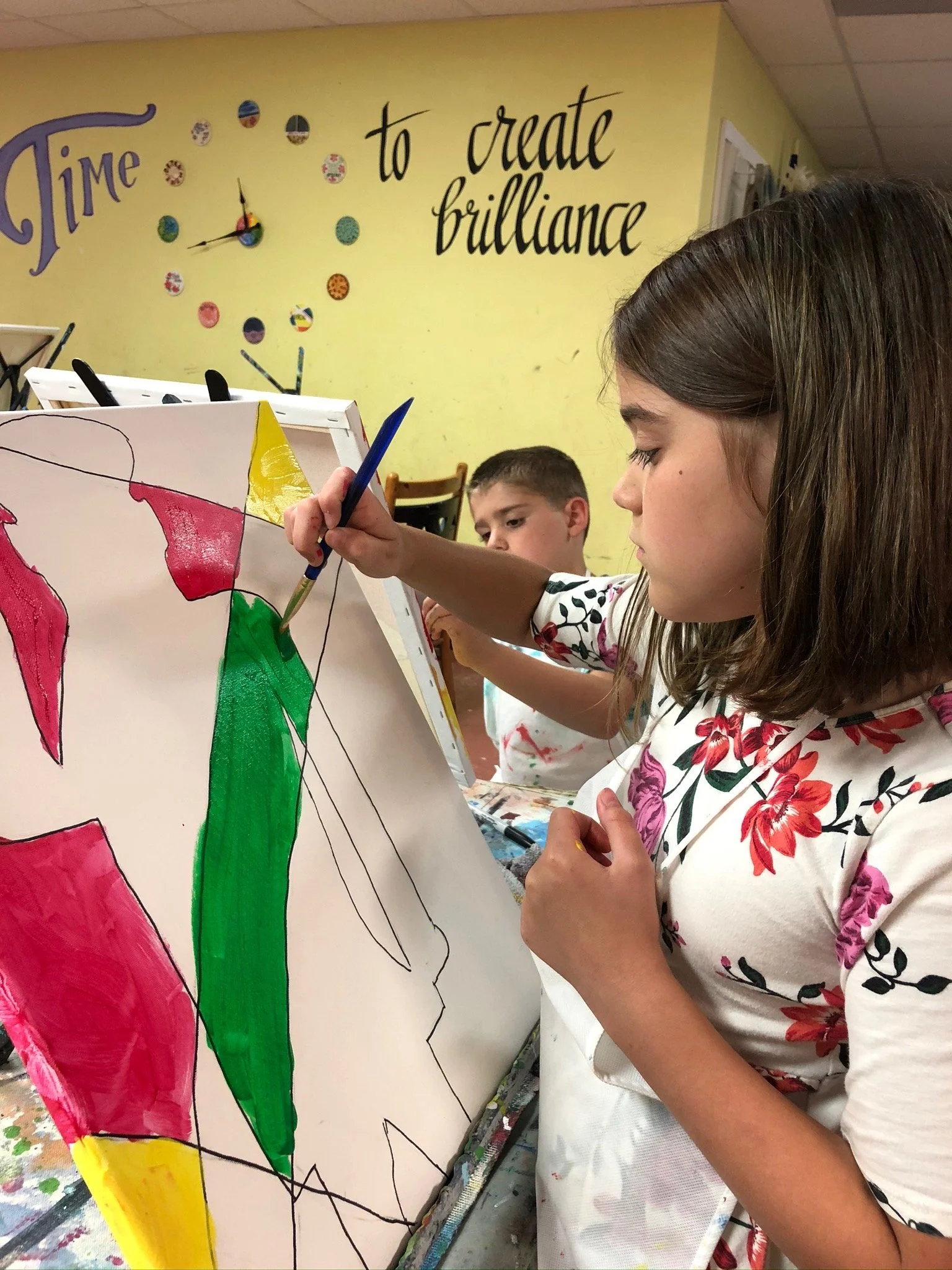 A girl in a floral shirt is painting abstract shapes with vibrant colors on a canvas in an art classroom, with another boy in the background observing.