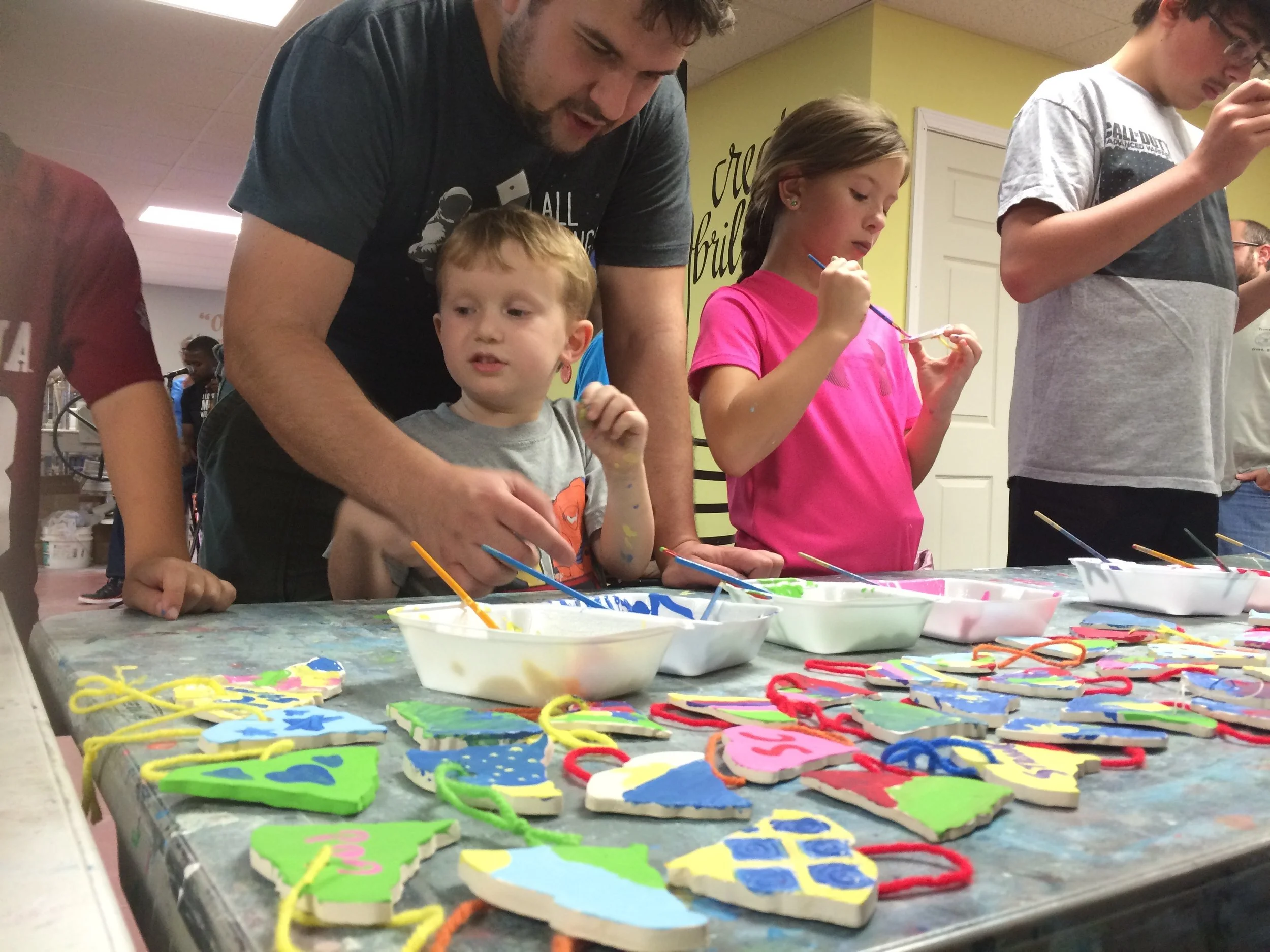 A group of children and an adult painting and decorating Christmas ornaments on a table. The ornaments are colorful and shaped like Christmas trees, with some having painted details and strings for hanging.