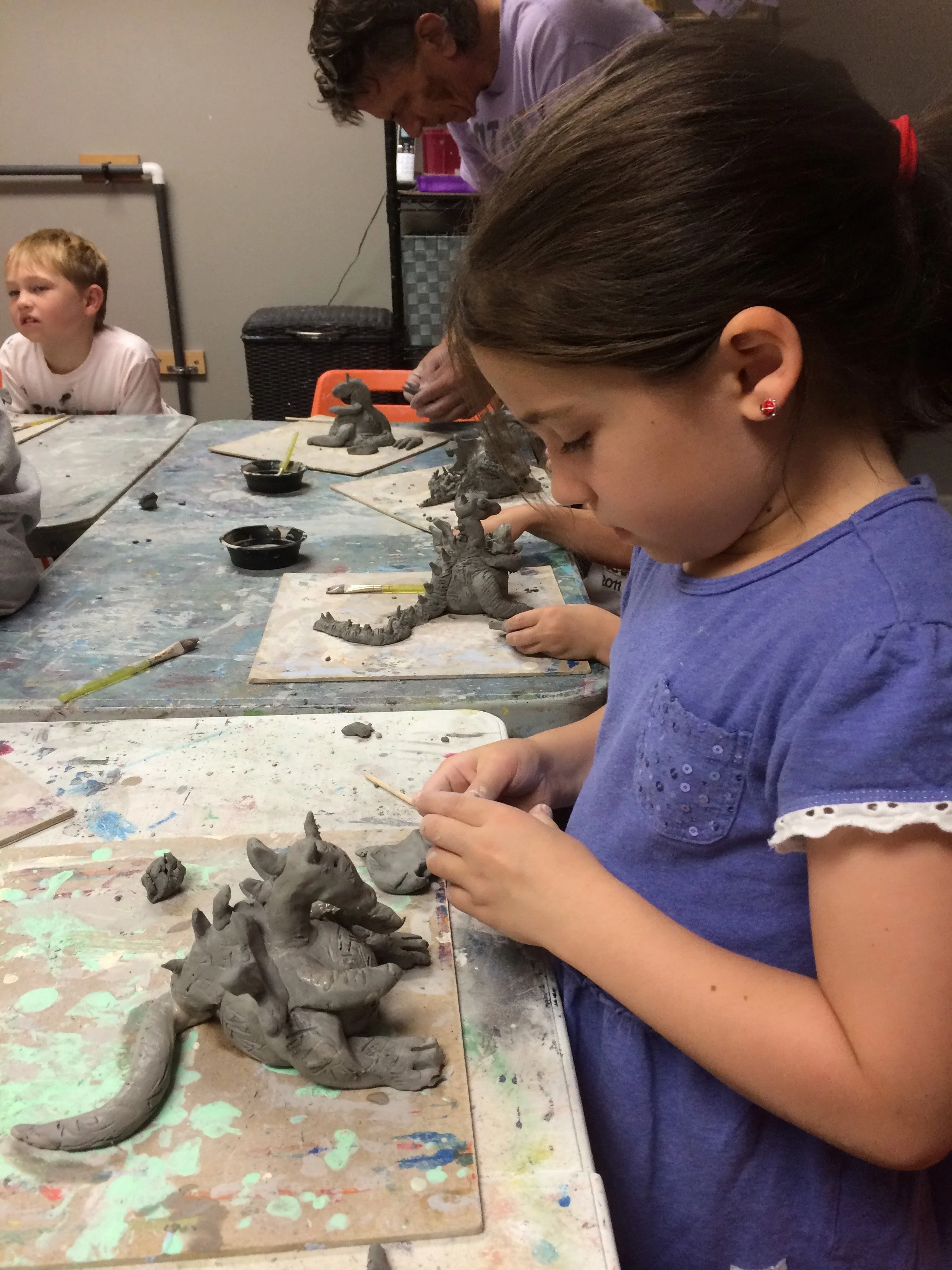 Young girl working on a clay sculpture of a creature resembling a dragon or lizard at an art class, with other children and an instructor also sculpting in the background.