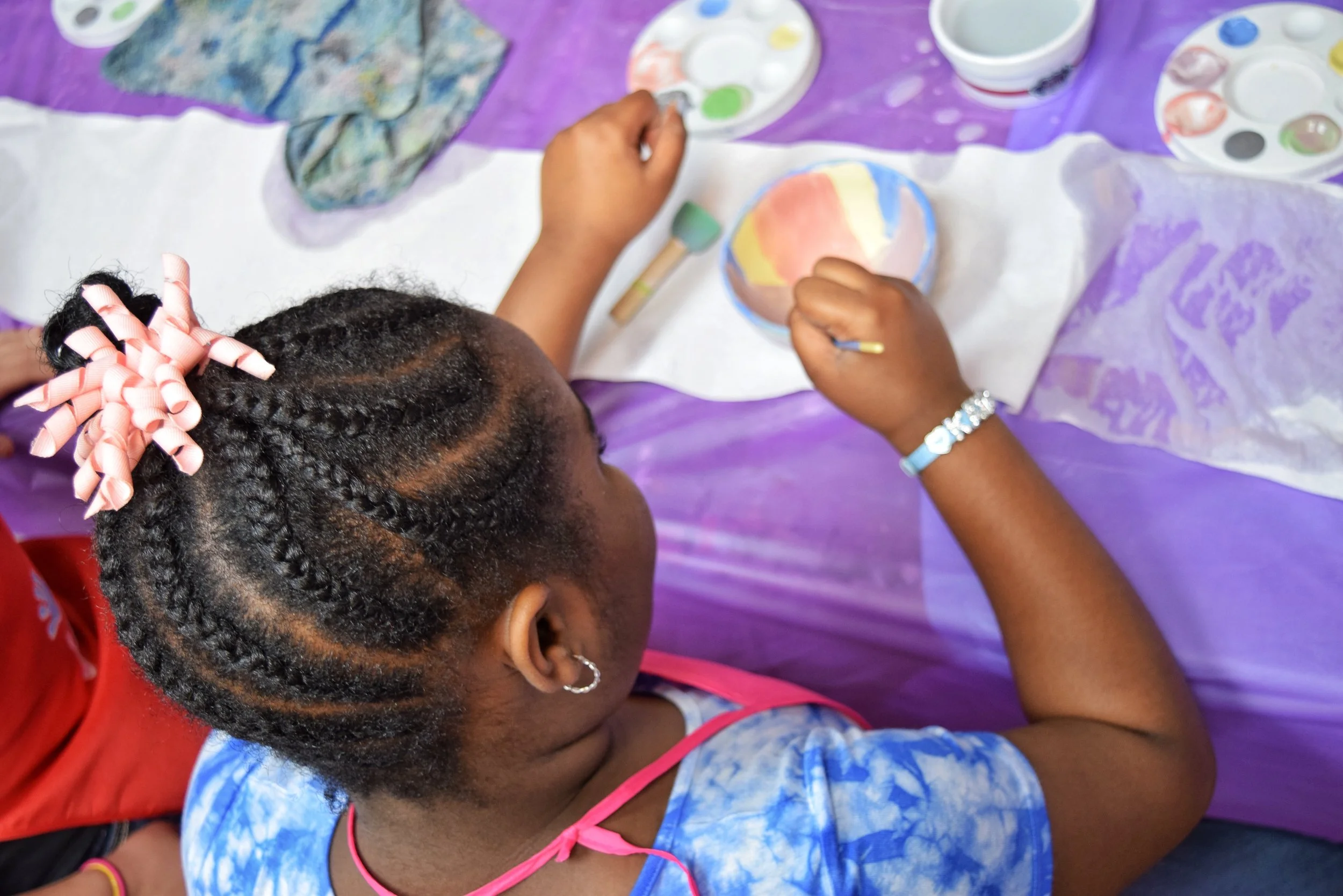 A young girl with braided hair and hair rollers painting on a ceramic bowl with watercolors at a craft table.