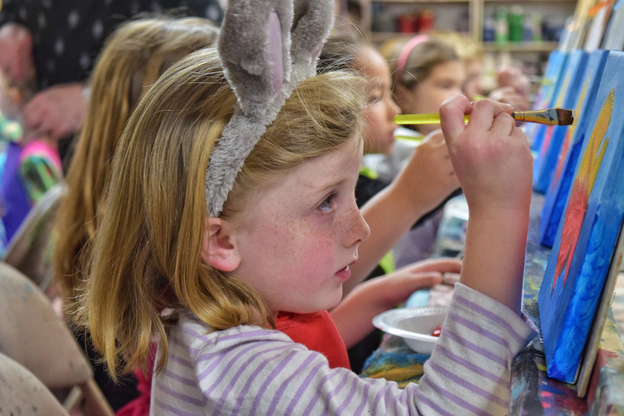 Children painting on canvases at an art class, girl in foreground wearing bunny ears headband.