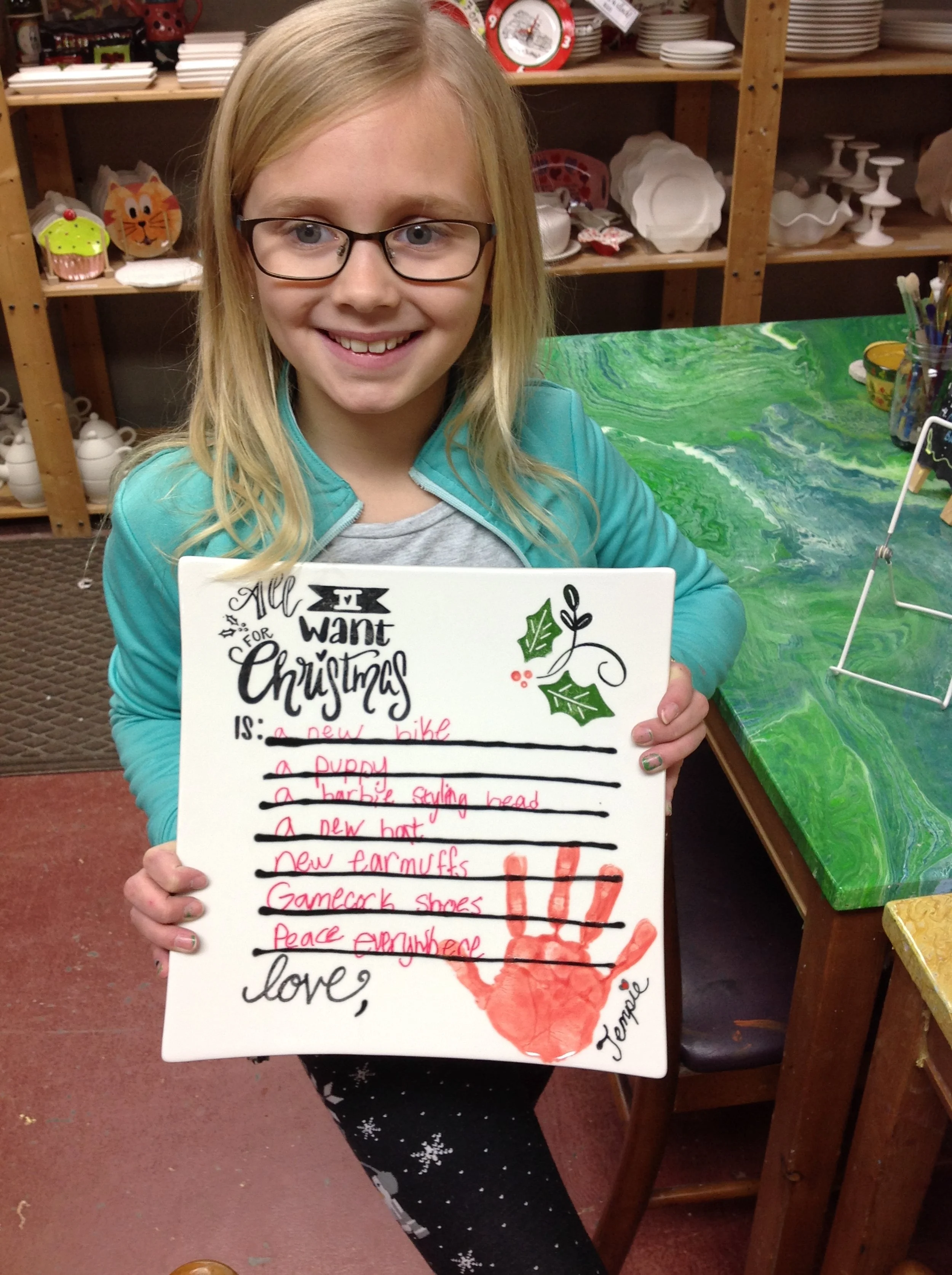 A young girl smiling and holding a whiteboard with a Christmas wish list written in pink marker. The list includes items like a new bike, puppy, and pajamas. The whiteboard has a red handprint with the name 'Jemelle' written below it, and the girl is