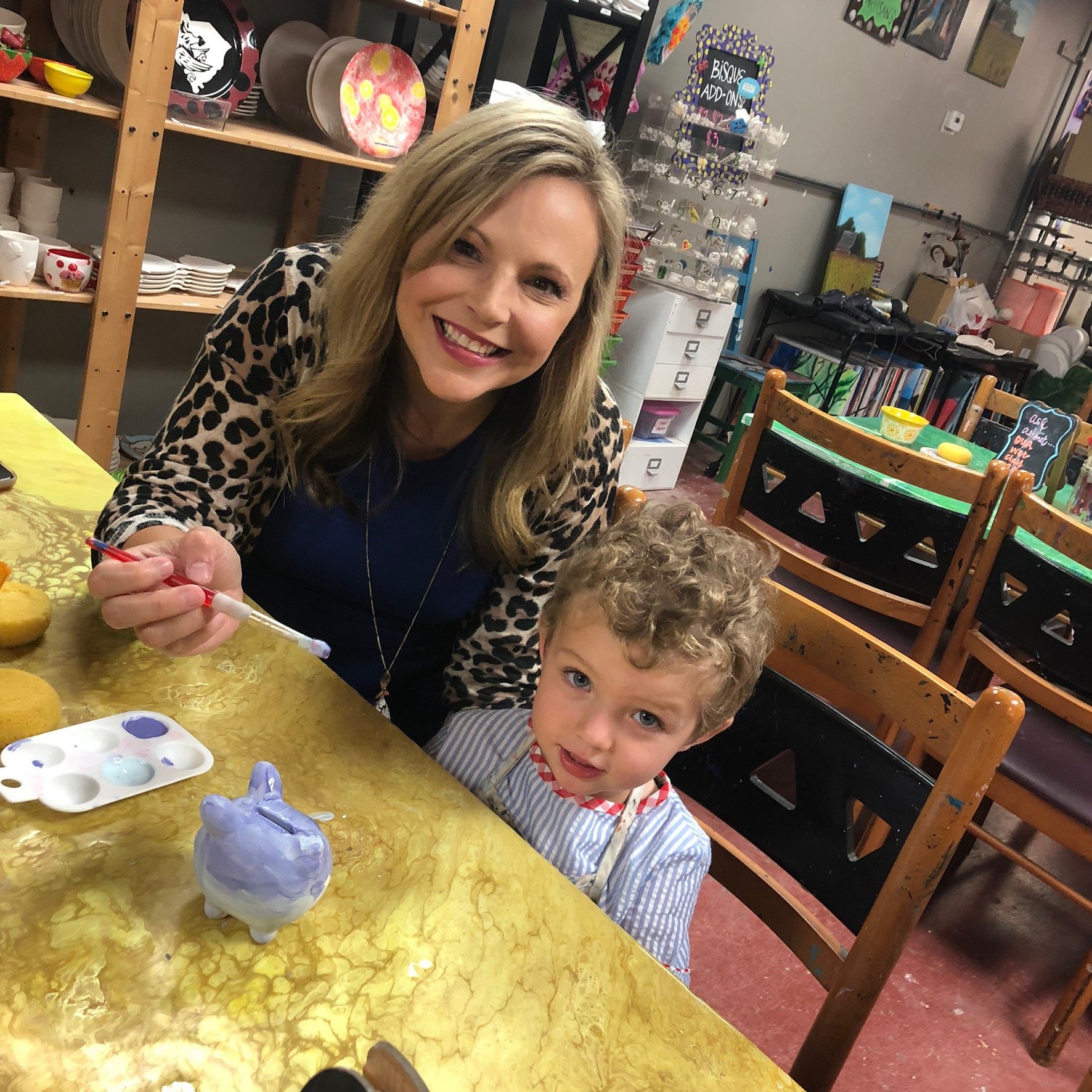 A woman and a young boy sitting at a table, engaging in a pottery painting activity. The woman is smiling and holding a paintbrush. The boy, with curly hair, is looking at the camera. The table has painted ceramic items and a paint palette, and the b