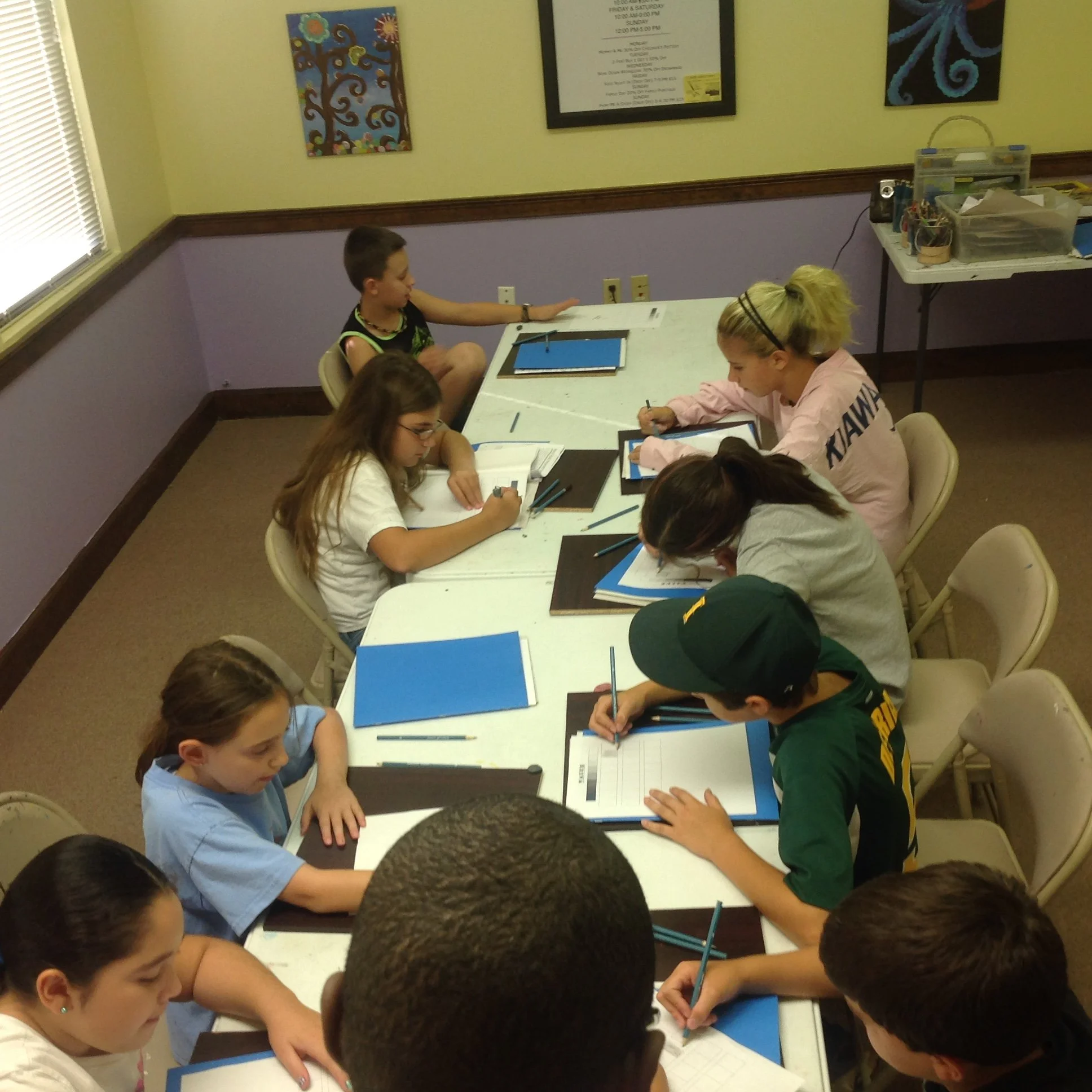 Children and young teens sitting around a white table, working on notebooks and worksheets with pens, in a classroom setting.