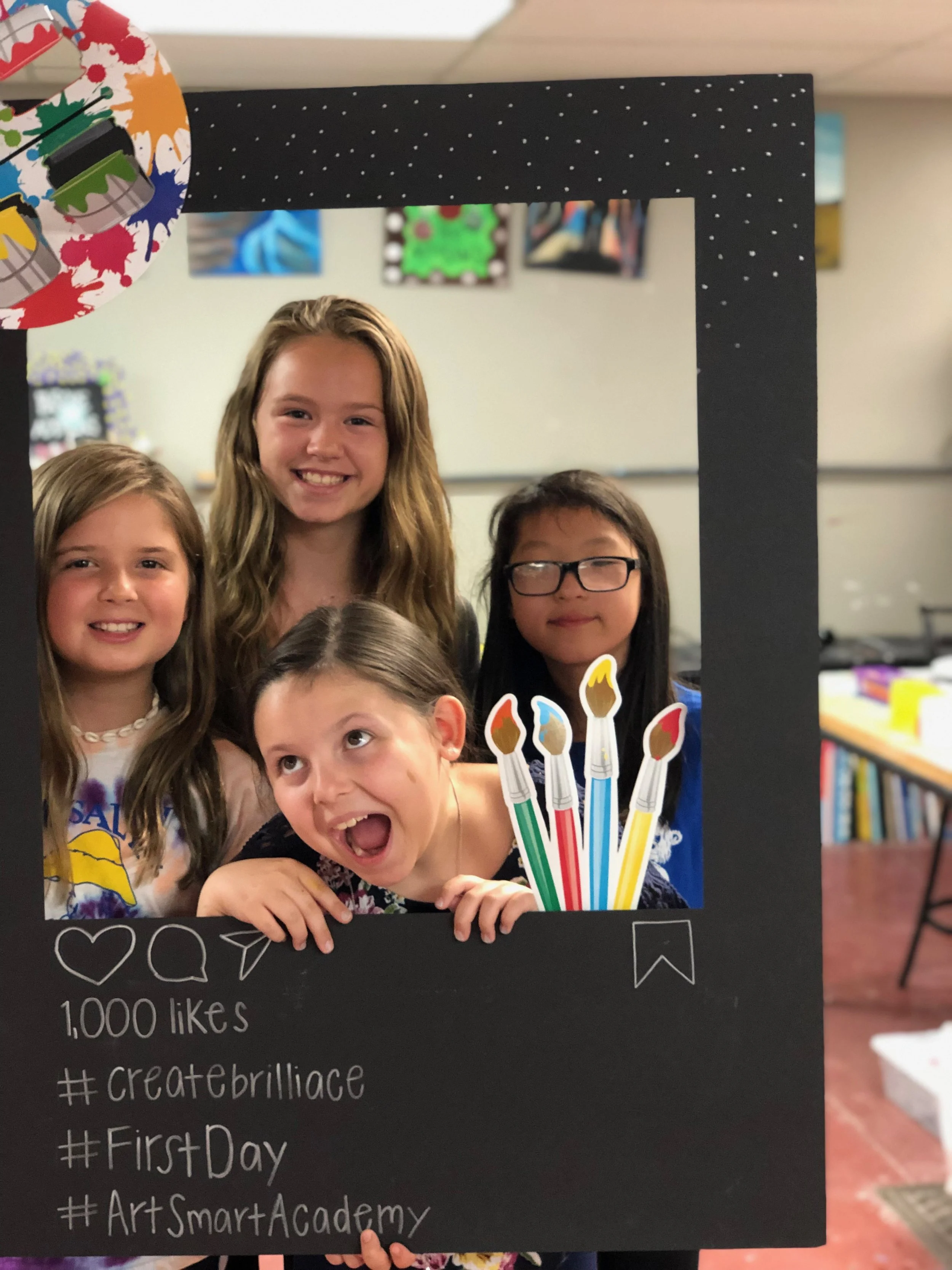 Group of five smiling children posing behind a black social media-style photo frame with birthday-themed decorations and chalk-writing, celebrating an event at an art school.