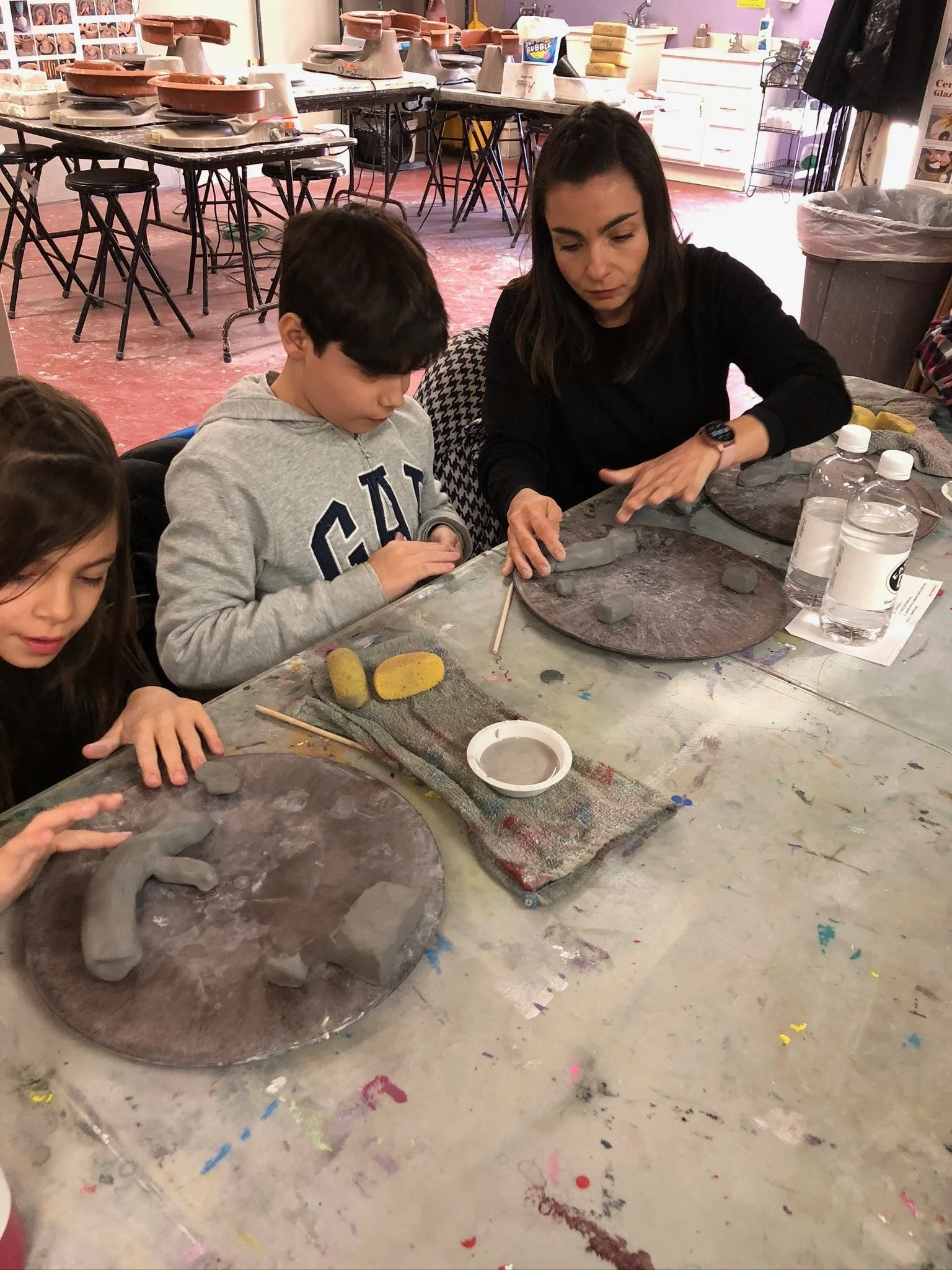 Children and an instructor working on clay projects at a pottery class, with pottery wheels in the background.