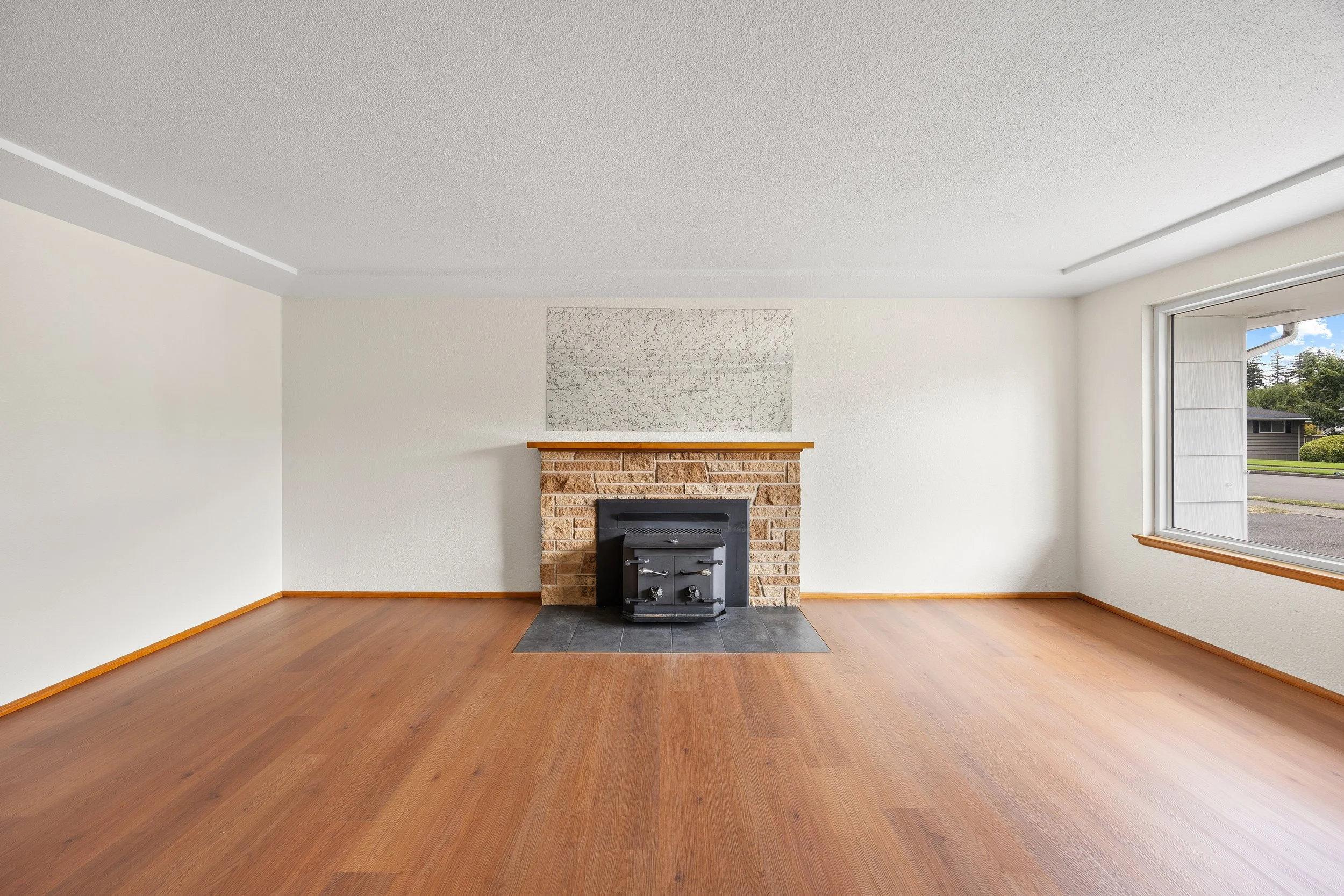 Empty living room with hardwood floors, a brick fireplace, and a large window facing the street. Real Estate photography in Lacey, Olympia, Tumwater, Seattle, Tacoma WA Drone Video 360 Tours
