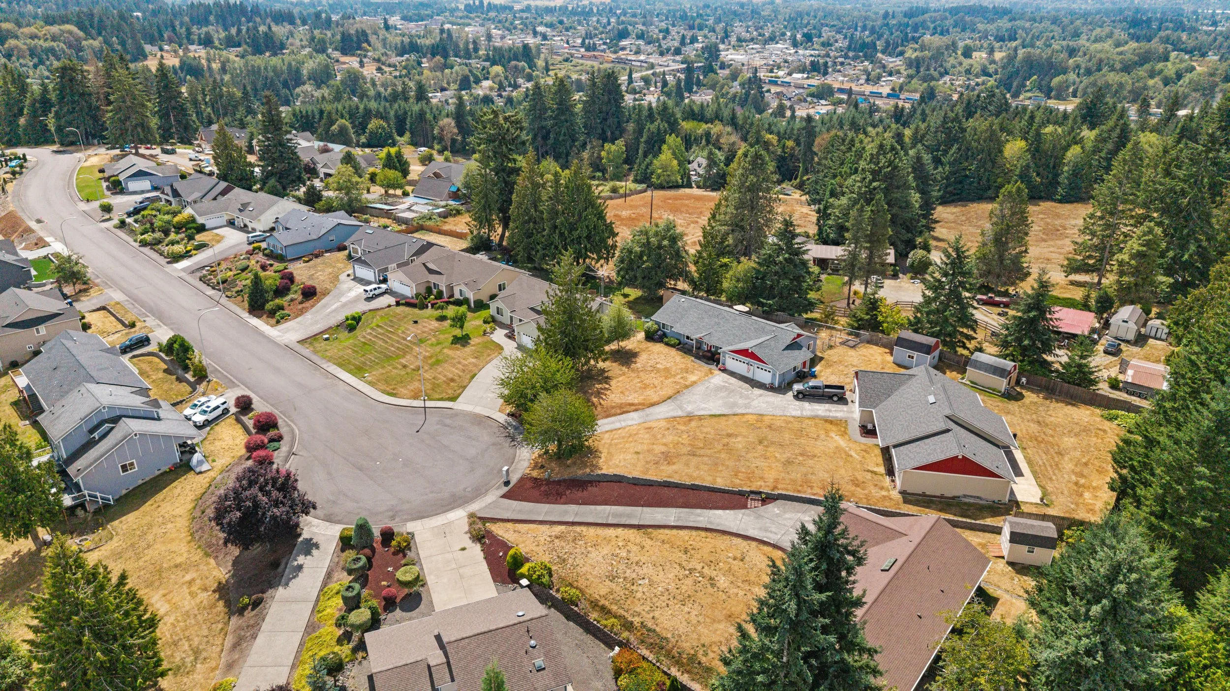 Aerial view of a suburban neighborhood with houses, lawns, trees, and streets, surrounded by a forested area. Real Estate Media; Photography, Drone, Video. Lacey, Olympia, Tumwater, Dupont, Centralia, Western WA
