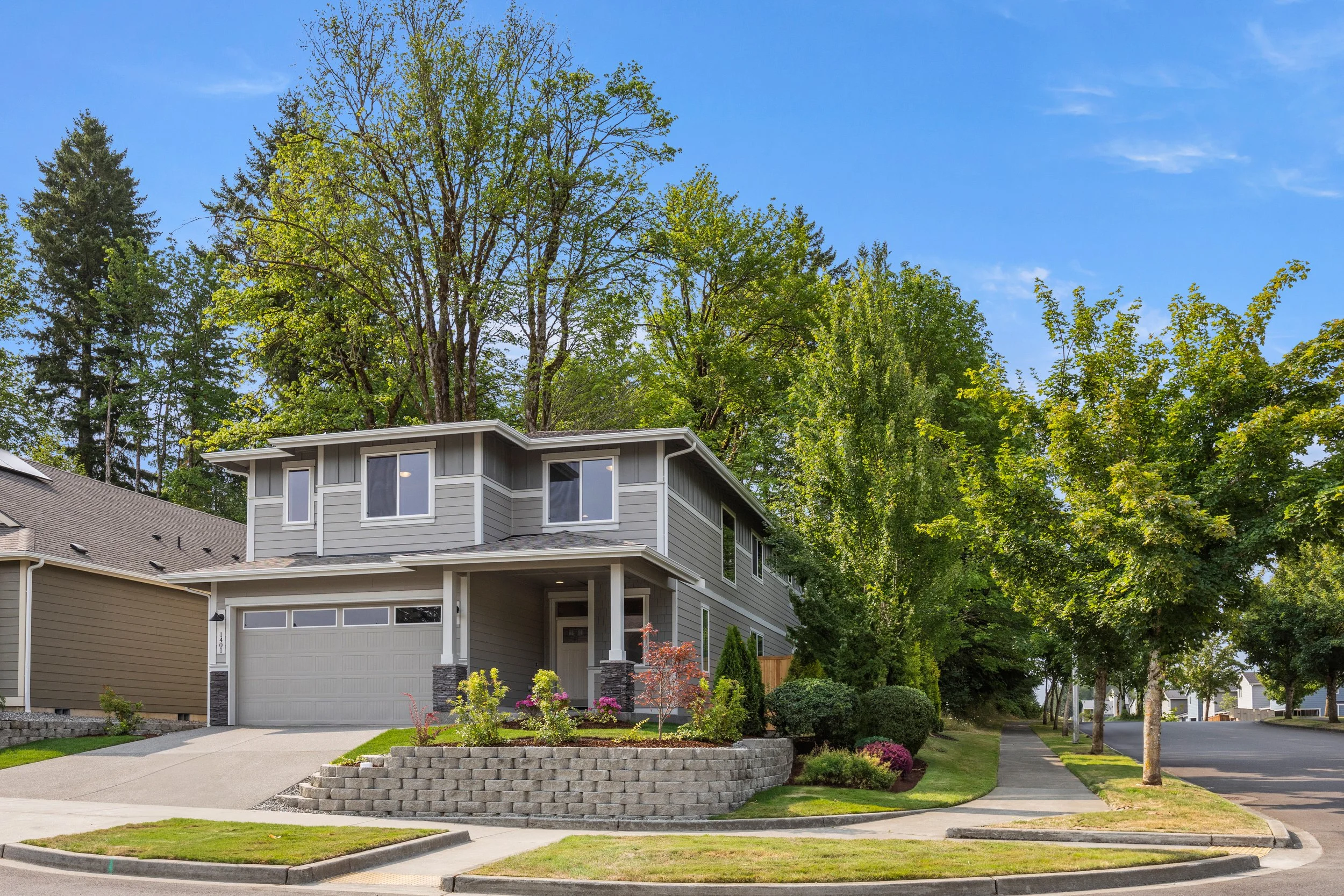 Modern two-story gray house with a garage and landscaped front yard, on a suburban street with trees and a clear blue sky. New Construction. Real Estate Media; Photography, Drone, Video. Lacey, Olympia, Tumwater, Dupont, Centralia, Western Washington
