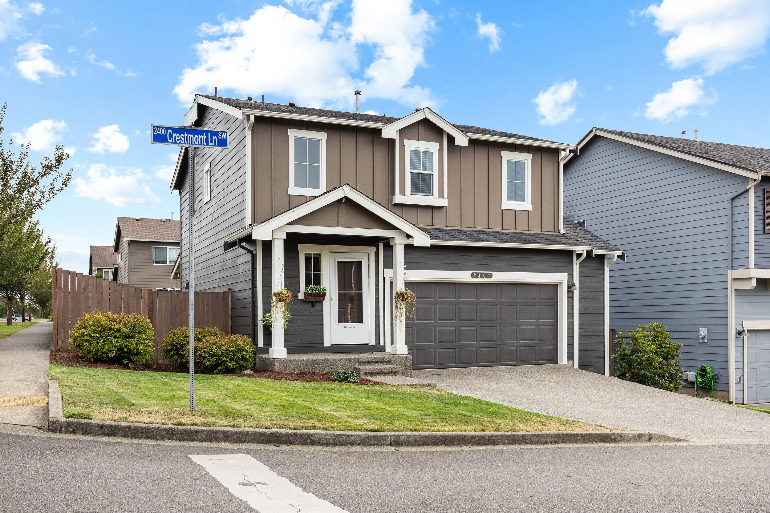 Two-story residential house with gray and brown exterior, front porch with columns, green lawn, driveway, and street sign in Lacey, Olympia, Tumwater, Washington, clear blue sky with scattered clouds. Real Estate Media; Photography, Drone, Video.