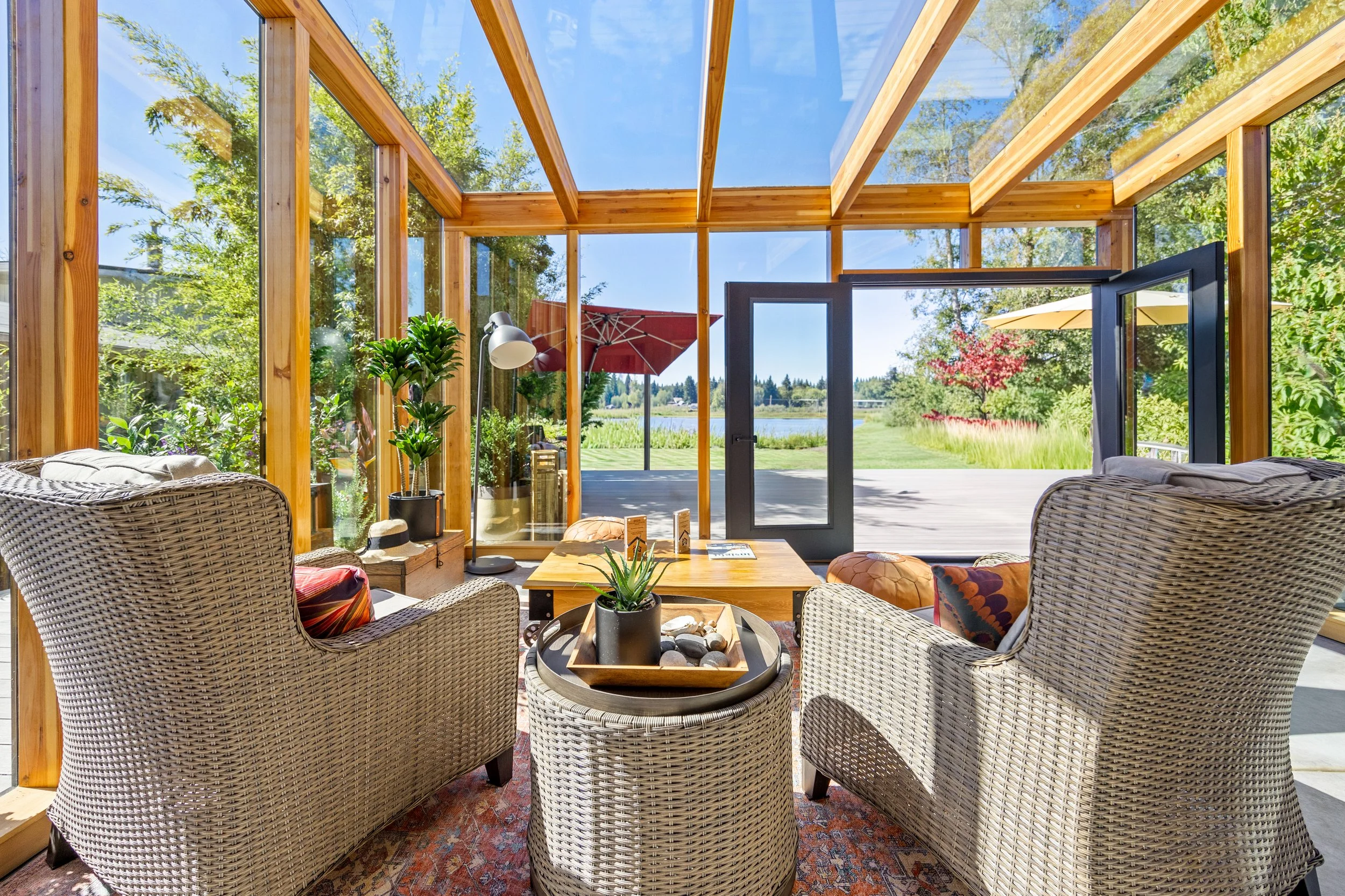 Sunroom with wicker armchairs, wooden coffee table, potted plants, and large glass windows showing a view of trees, grass, and a lake. Real Estate photography in Lacey, Olympia, Tumwater, Seattle, Tacoma WA Drone Video 360 Tours