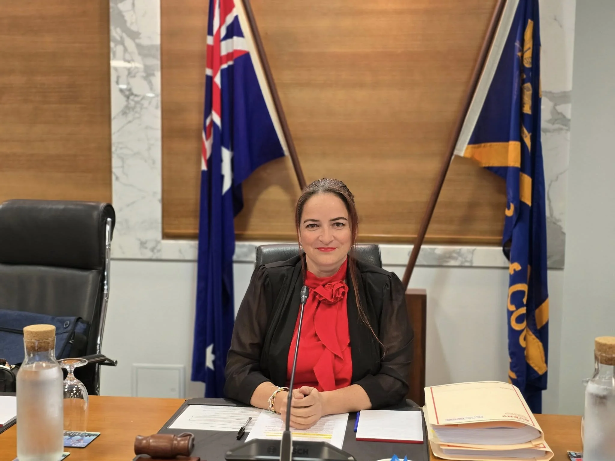 A woman sitting at a conference table with flags in the background, wearing a black blazer and red blouse, smiling at the camera.
