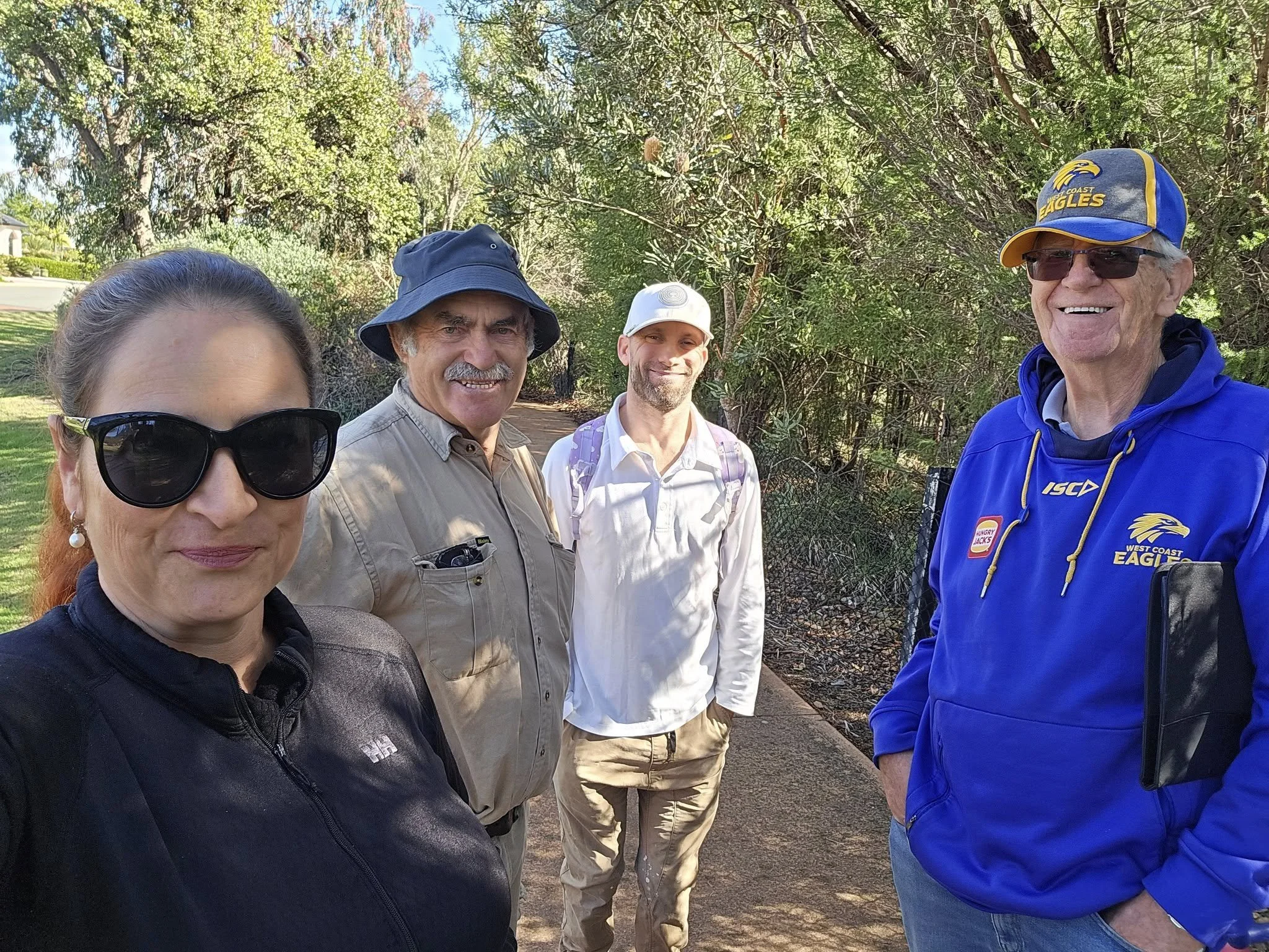 Four people smiling and standing outdoors on a sunny day, with trees and a fence in the background.