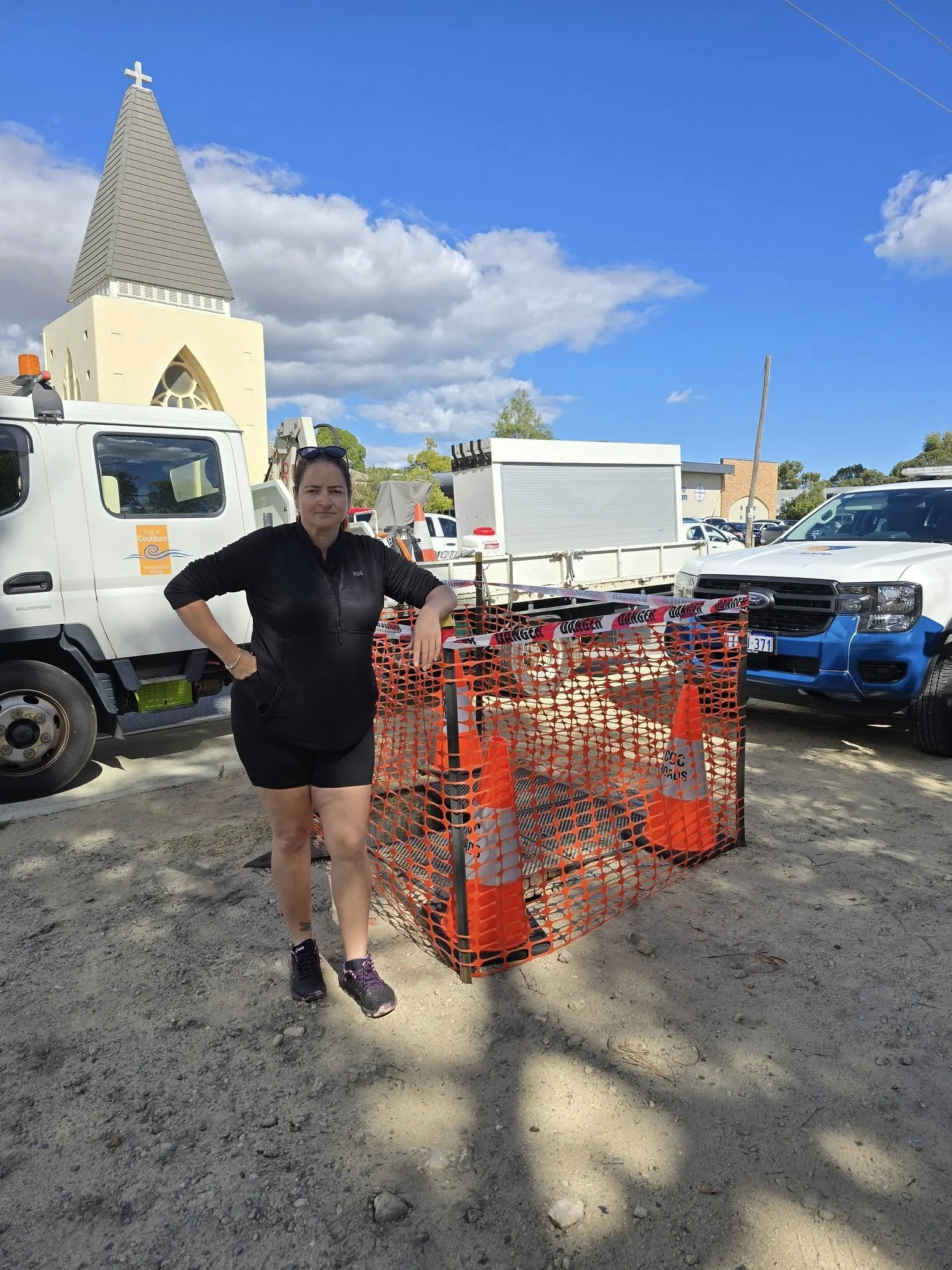 A woman in black athletic clothing stands next to a fenced safety area containing orange traffic cones on a dirt parking lot with trucks and a church in the background under a partly cloudy sky.