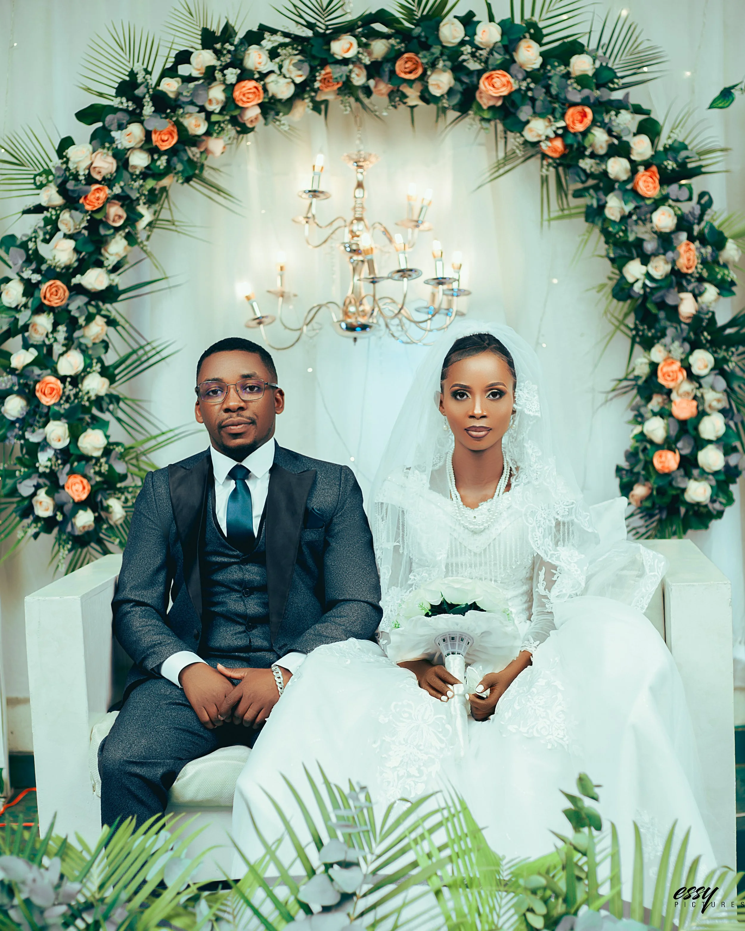 A bride and groom sitting on a white couch at their wedding. The bride is wearing a white wedding gown with lace details, holding a bouquet of white roses and wearing a pearl necklace and lace veil. The groom is dressed in a dark suit with a tie. The