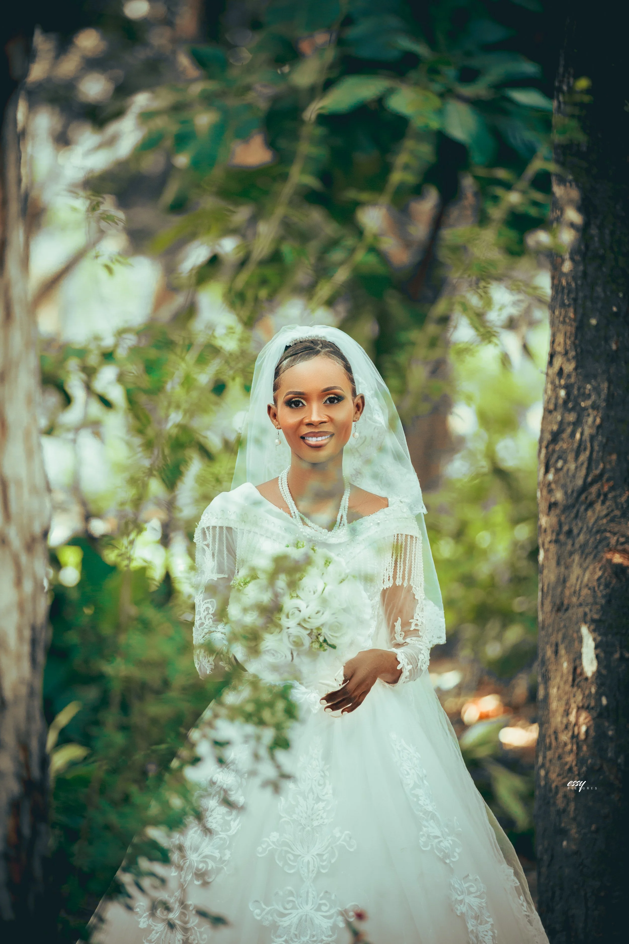 A bride in a white wedding dress holding a bouquet of white roses, standing outdoors among trees and greenery.