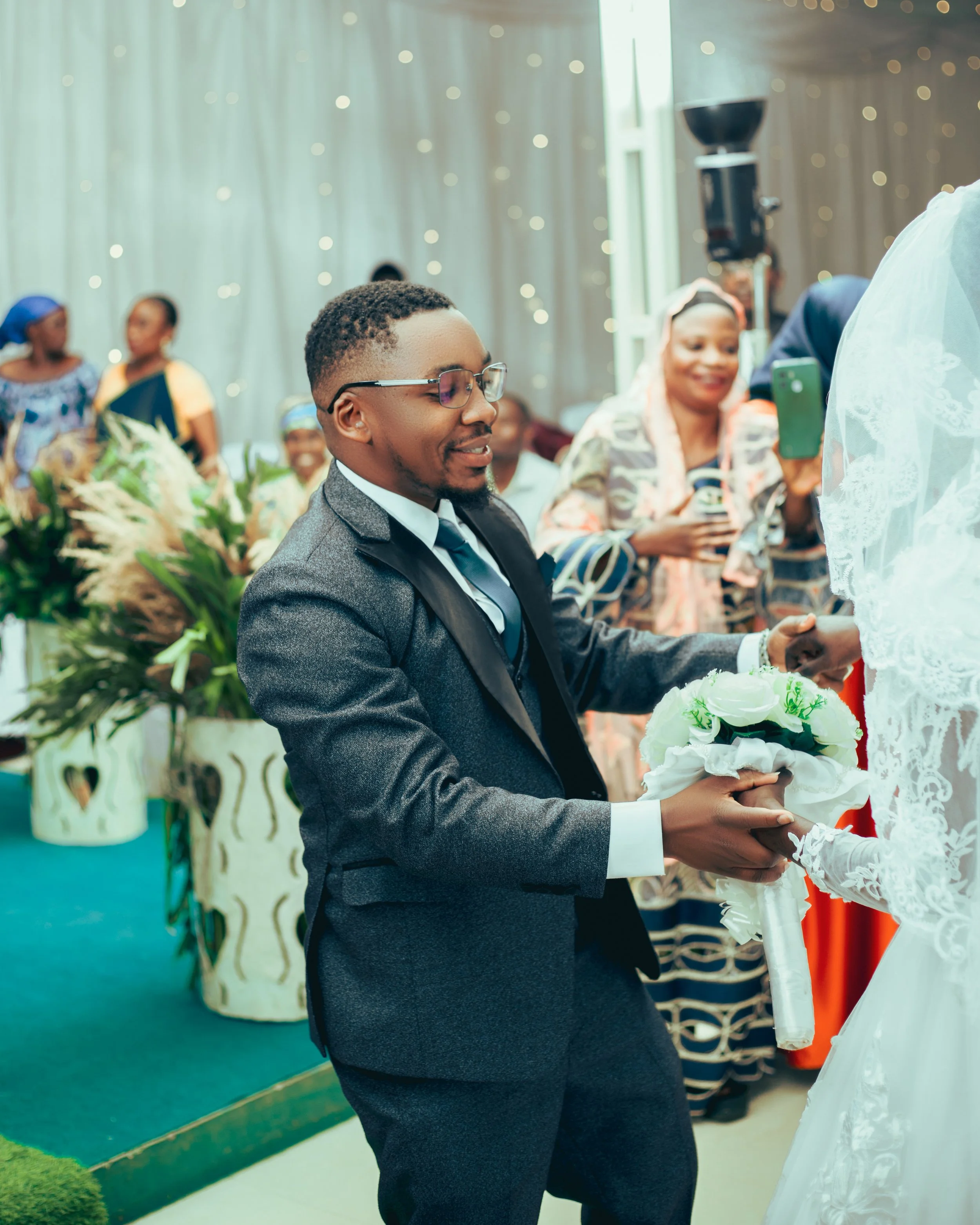 A groom in glasses and a dark gray suit holding a bouquet while shaking hands with a bride in a white lace dress at a wedding ceremony. Wedding guests, including a woman taking a photo, are visible in the background.