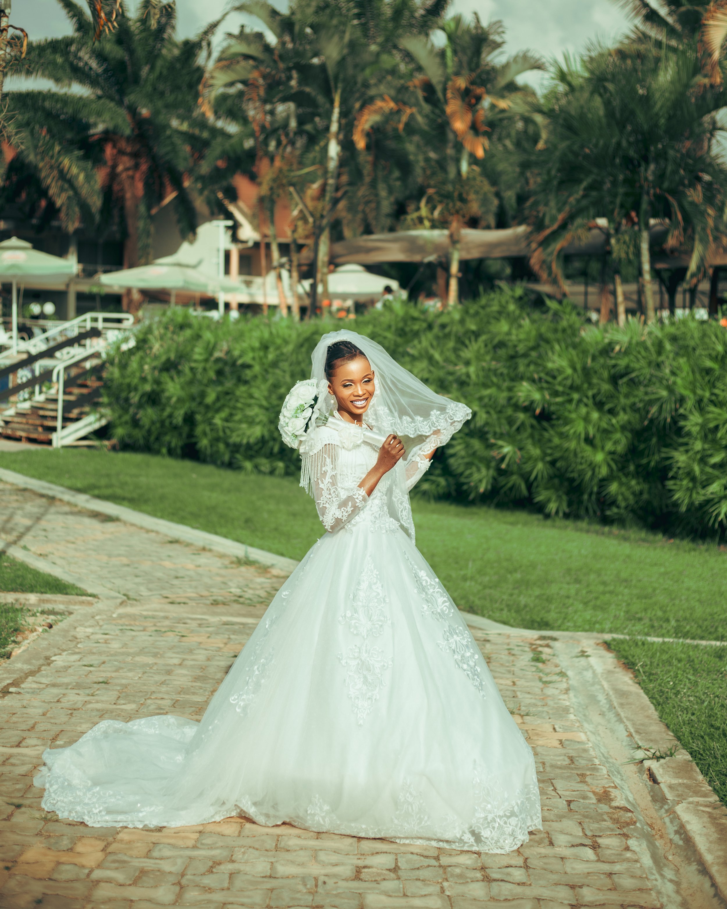 A woman in a white wedding dress with lace details is smiling and holding her veil outside on a brick pathway, with greenery and palm trees in the background.