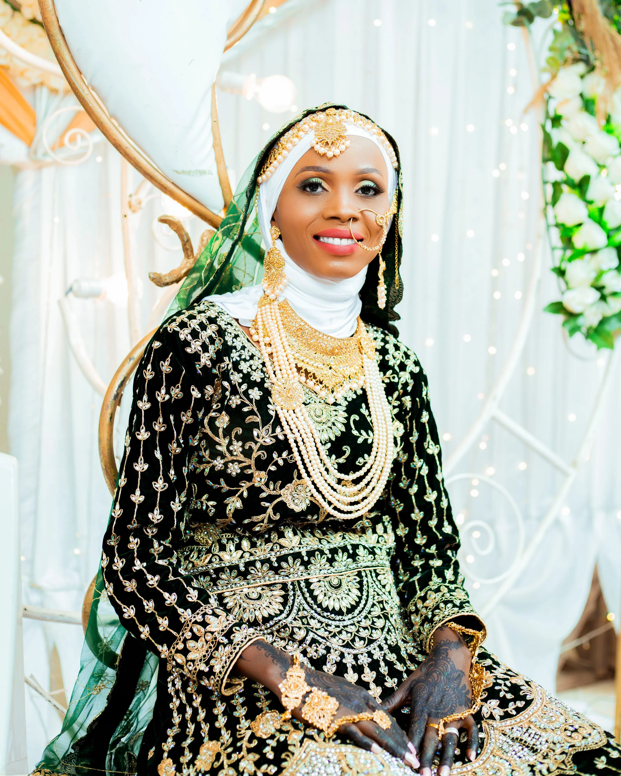 A woman dressed in traditional black and gold African attire, decorated with elaborate gold jewelry, sitting with a smile at a festive event.