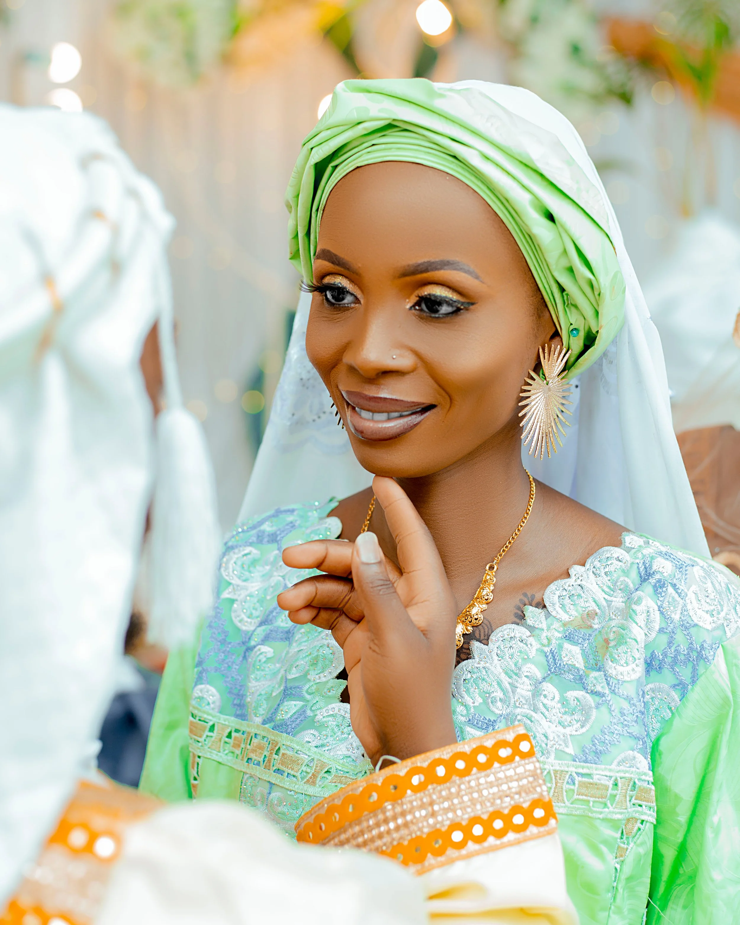 A woman with dark skin dressed in traditional attire, wearing a green headwrap and jewelry, is smiling and touching her chin with her right hand at a celebration or special event.