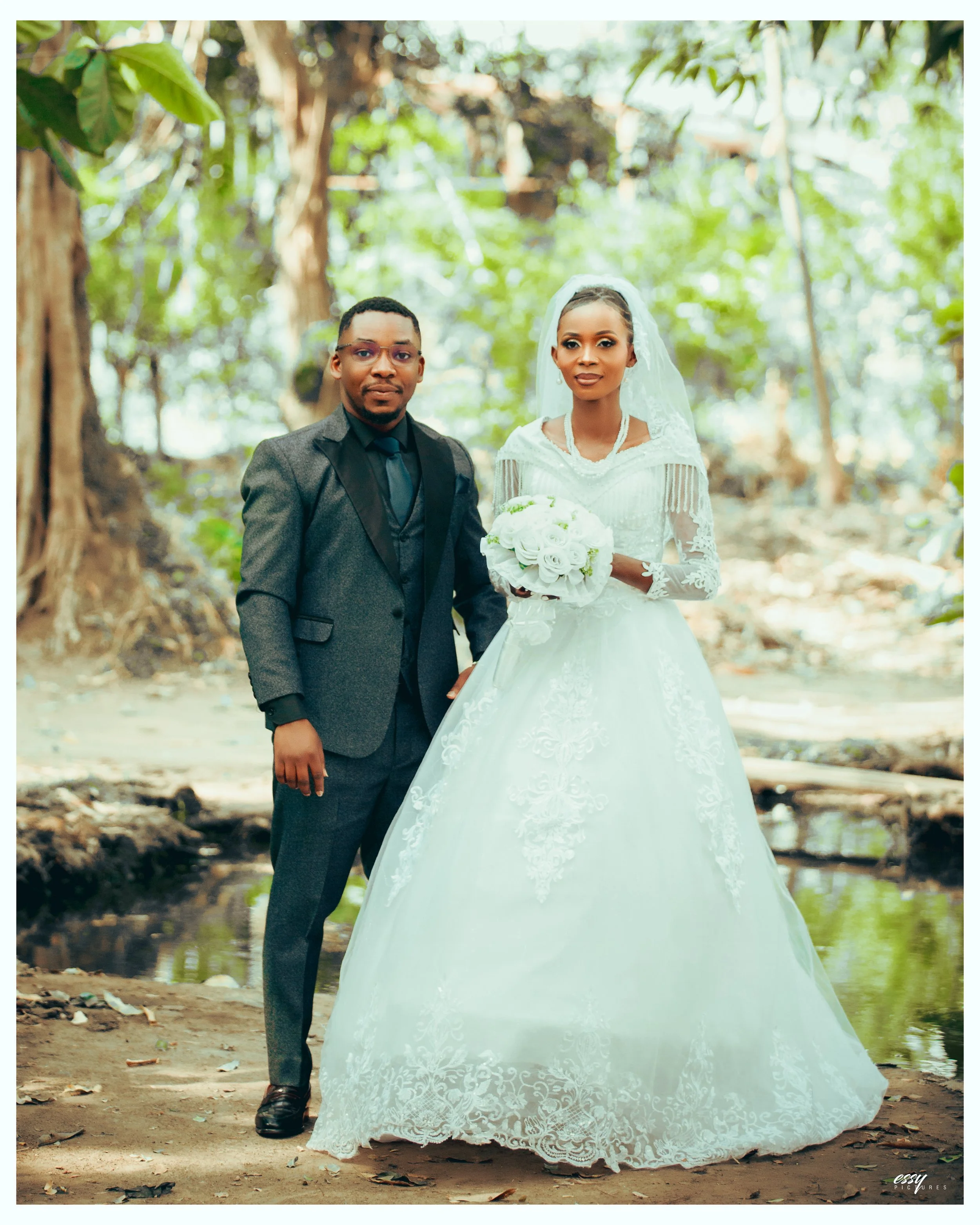 A bride and groom stand outdoors in a wooded area, with the bride wearing a white wedding gown and holding a bouquet of white roses, and the groom in a dark suit with a shirt and tie.