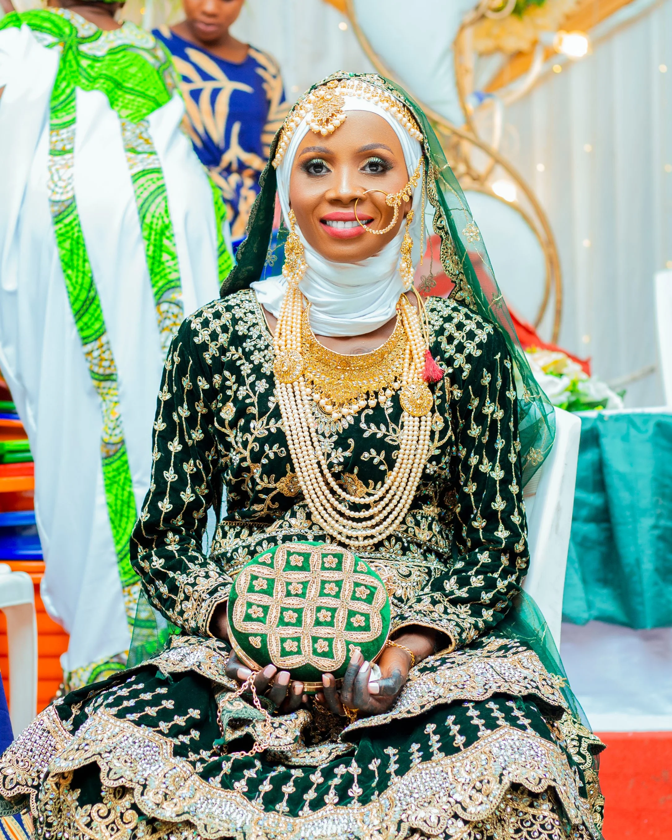 Woman in traditional Nigerian attire adorned with gold jewelry, wearing a white headscarf, holding a green and gold embroidered clutch, seated at a celebration.