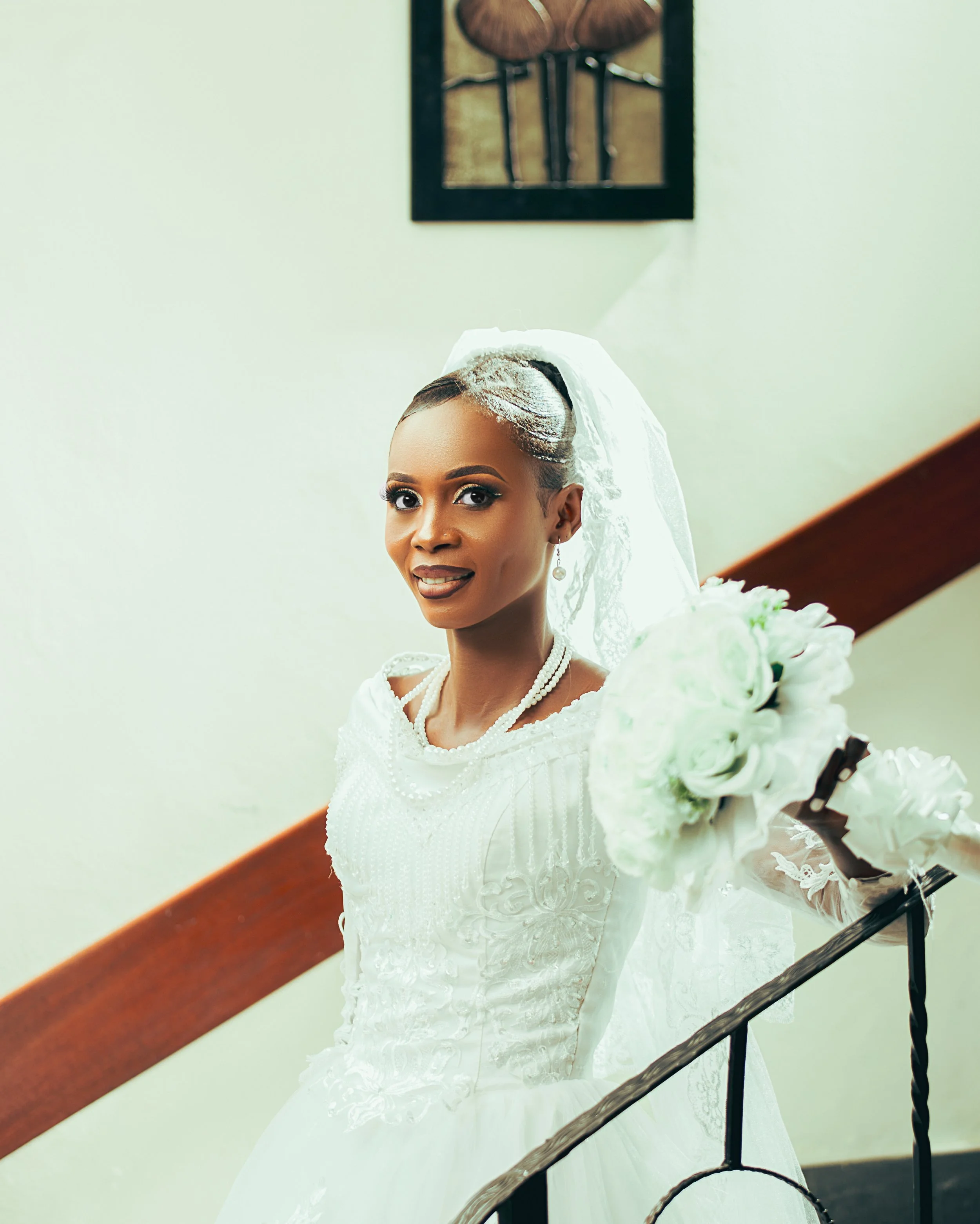 A bride with dark hair styled in an updo, wearing a white wedding dress and veil, holding a bouquet of white roses, standing on stairs indoors.