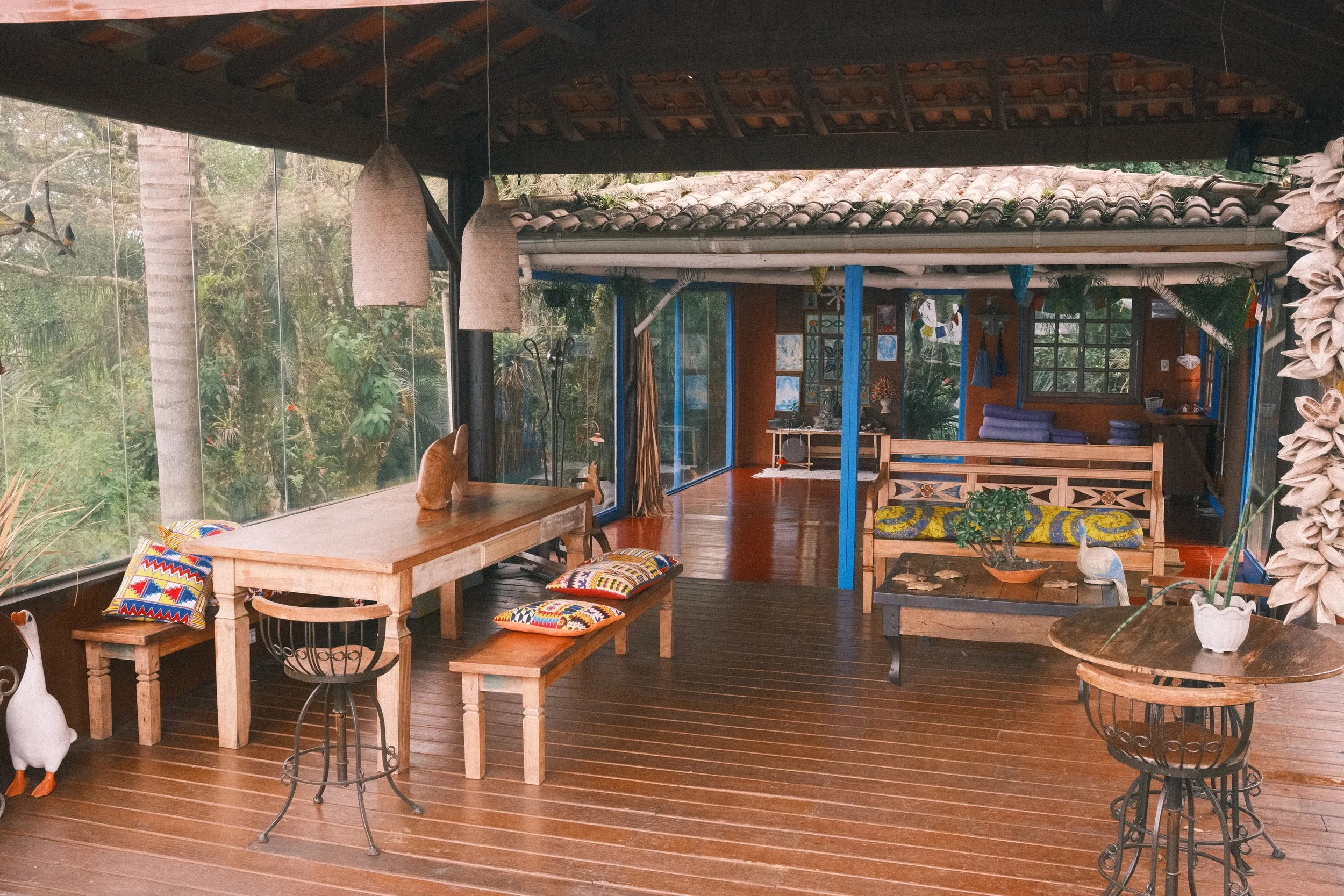 Indoor living space with wooden furniture, colorful cushions, and large windows showing greenery outside.