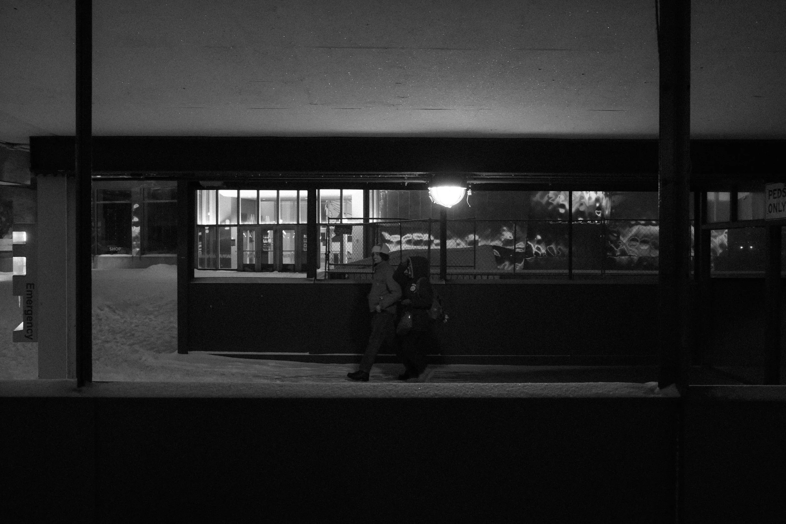 Two people walking past a covered outdoor area at night, with a winter landscape and snow on the ground, and a partially lit window in the background.