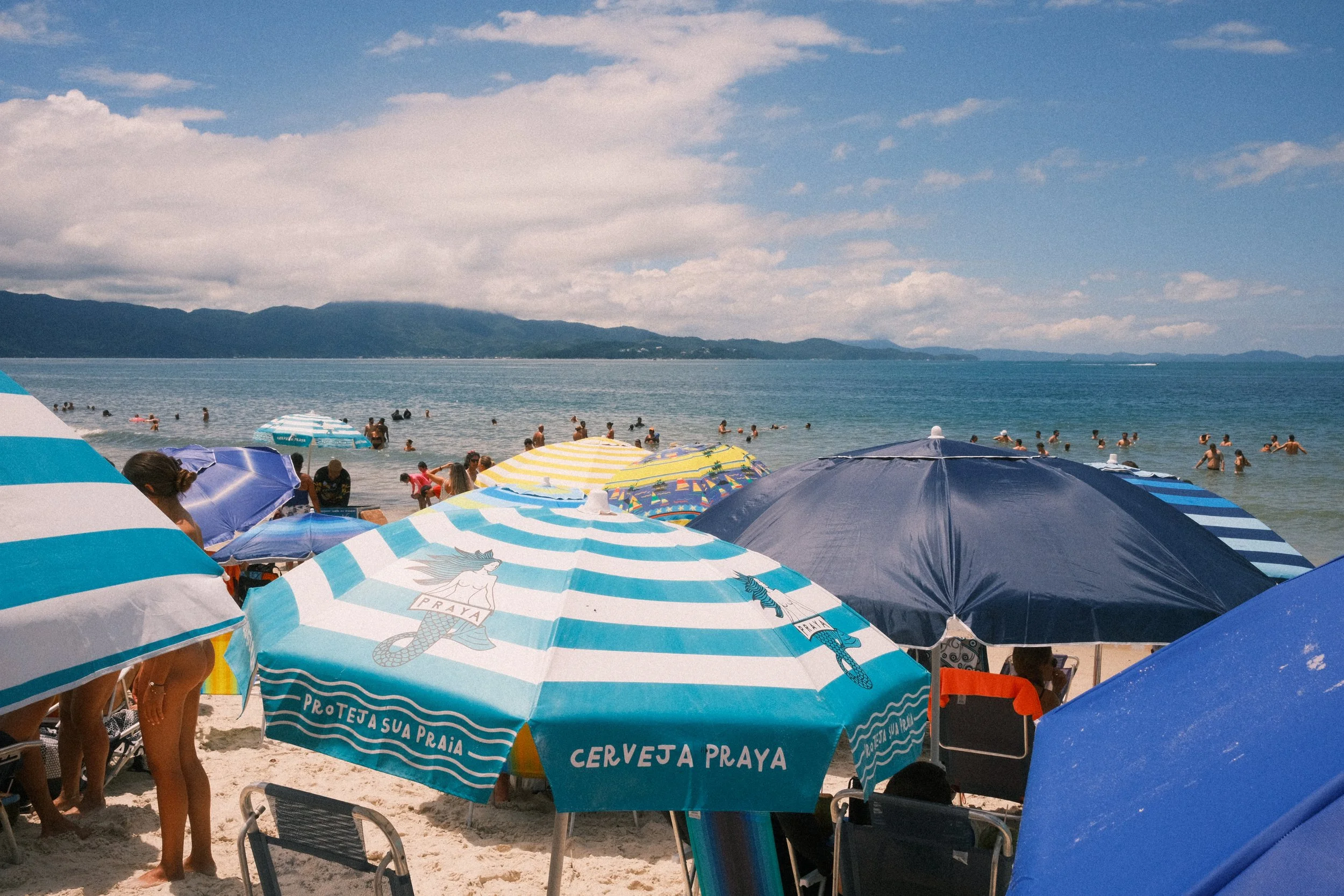 A crowded beach scene with colorful striped and solid umbrellas, people swimming and relaxing in the water, and a mountain range in the distance under a partly cloudy sky.