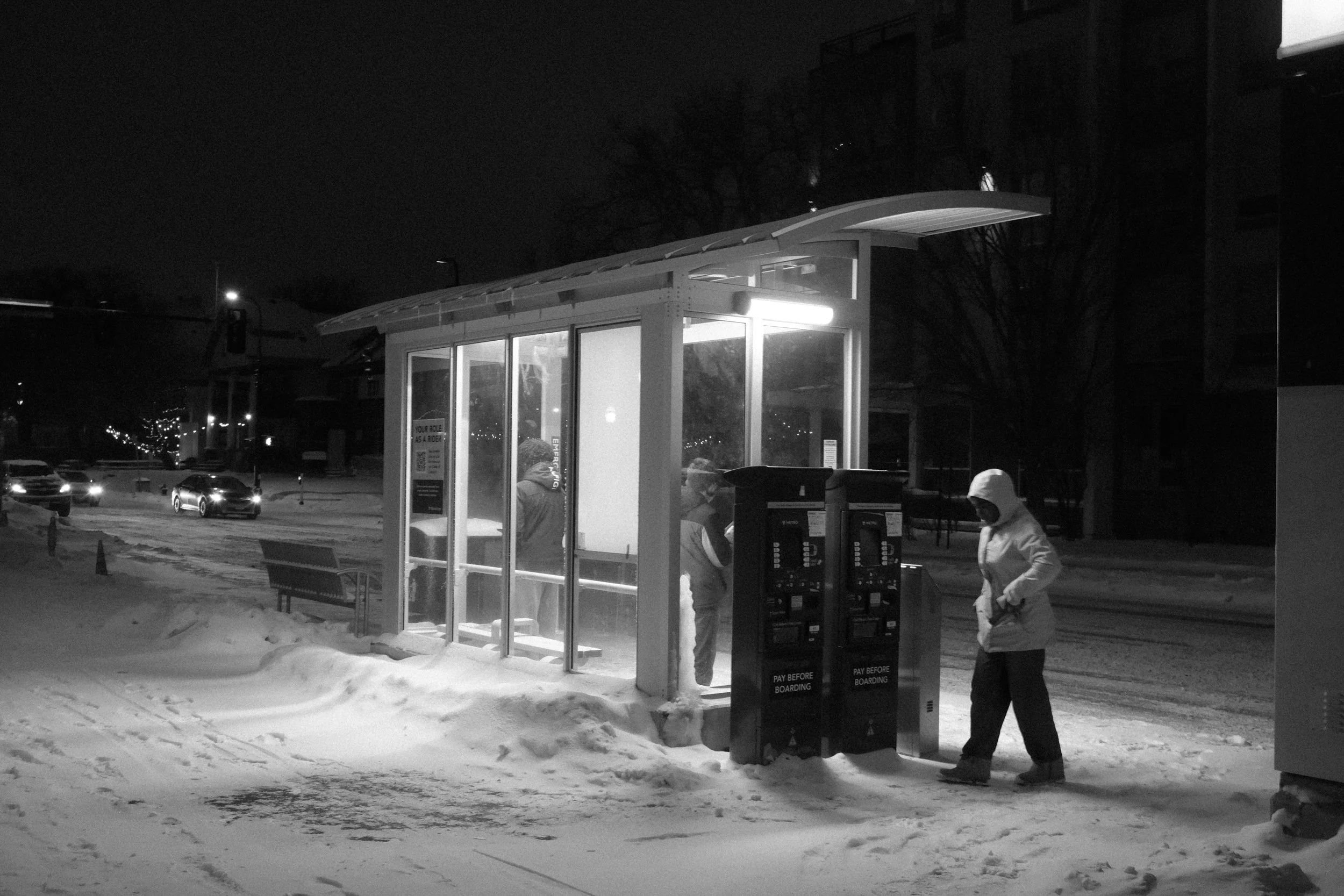 Nighttime scene of a snow-covered bus stop shelter with people inside and outside, and two ticket machines in front of the shelter.