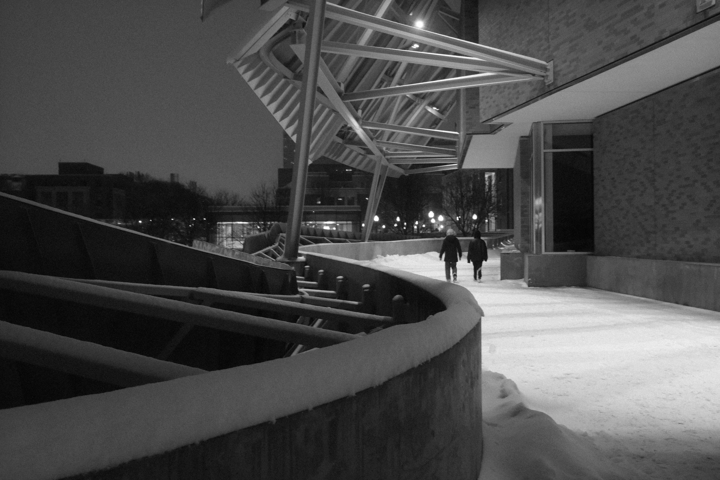 Nighttime black-and-white photo of two people walking on a snow-covered sidewalk beside a modern building with a large, angular metal sculpture.