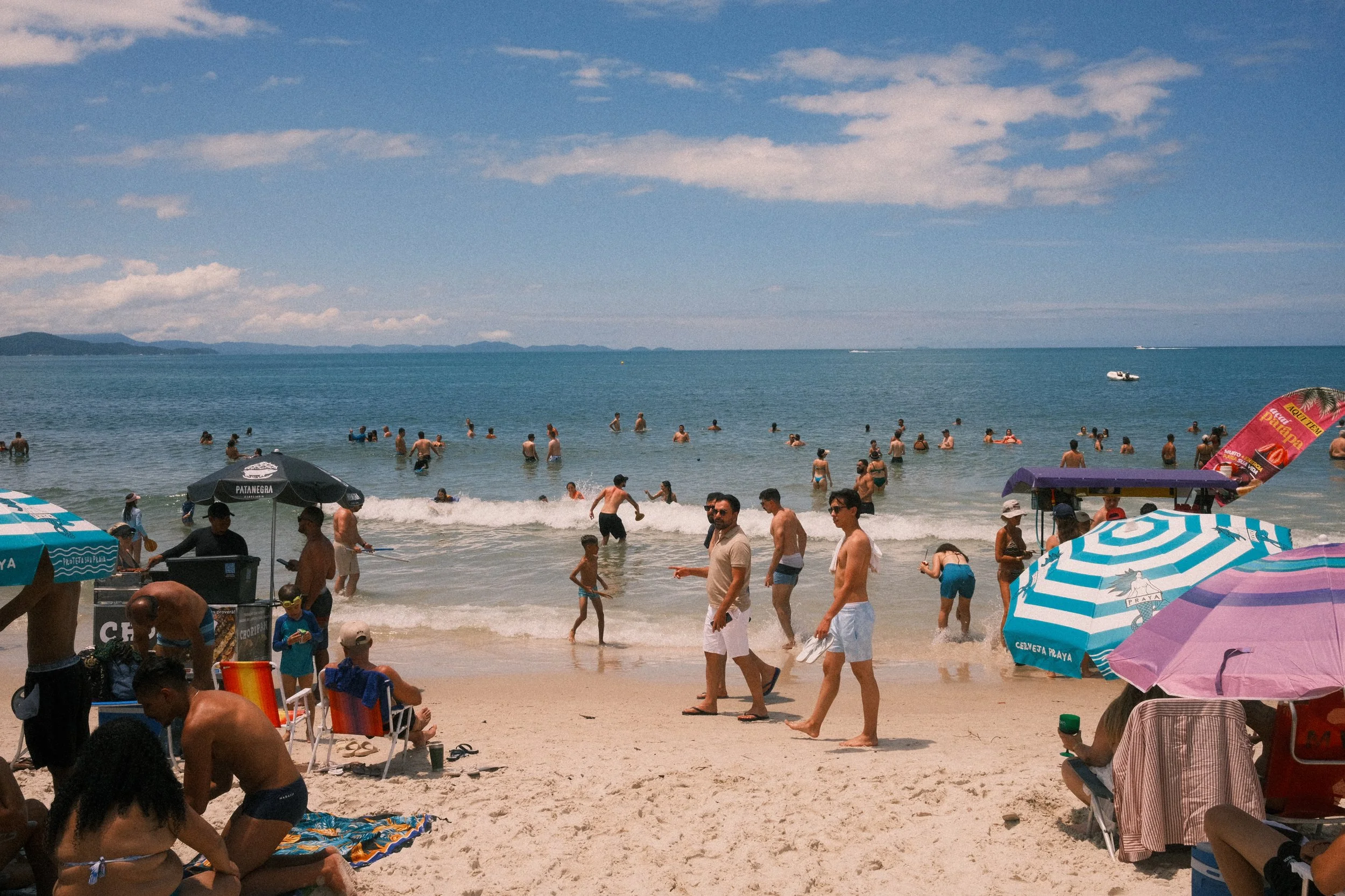 Busy beach scene with people swimming in the ocean and relaxing under umbrellas on the sandy shore on a sunny day.