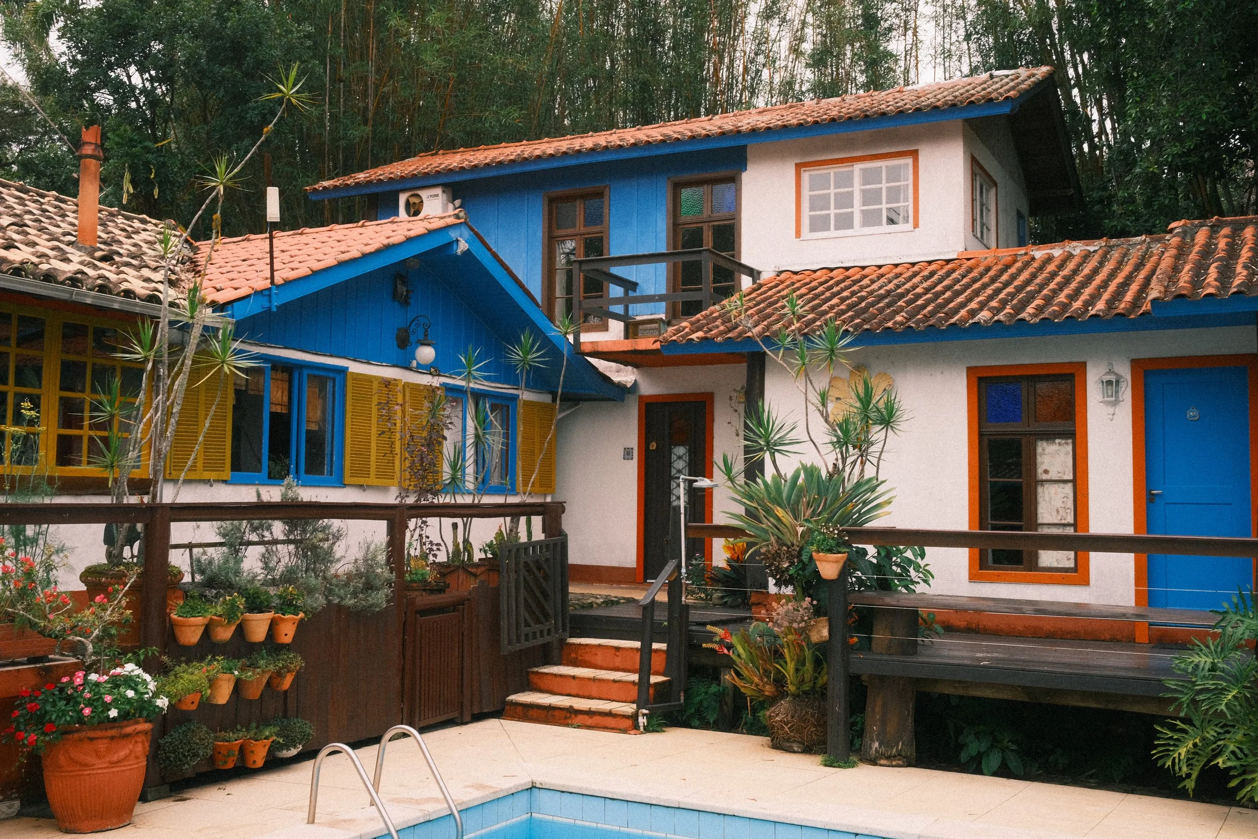 Colorful two-story house with blue, white, and yellow walls, surrounded by potted plants and trees, with a swimming pool in the foreground.