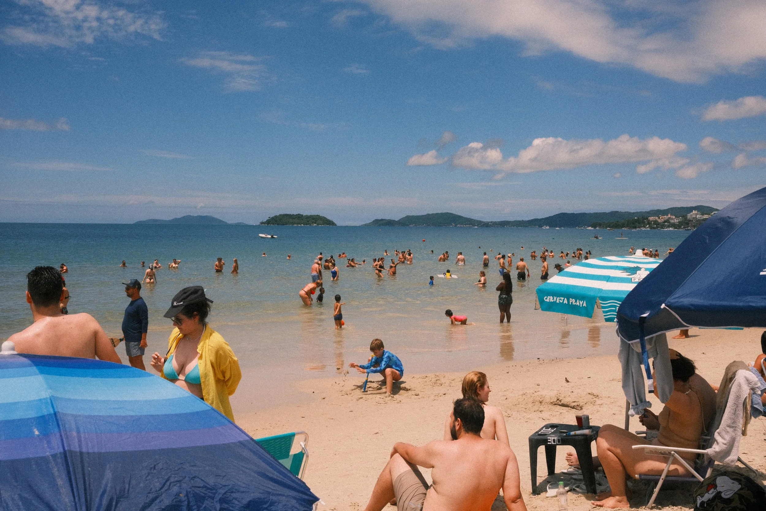 People swimming and relaxing on a crowded beach with umbrellas, and a view of the water and distant islands under a partly cloudy sky.