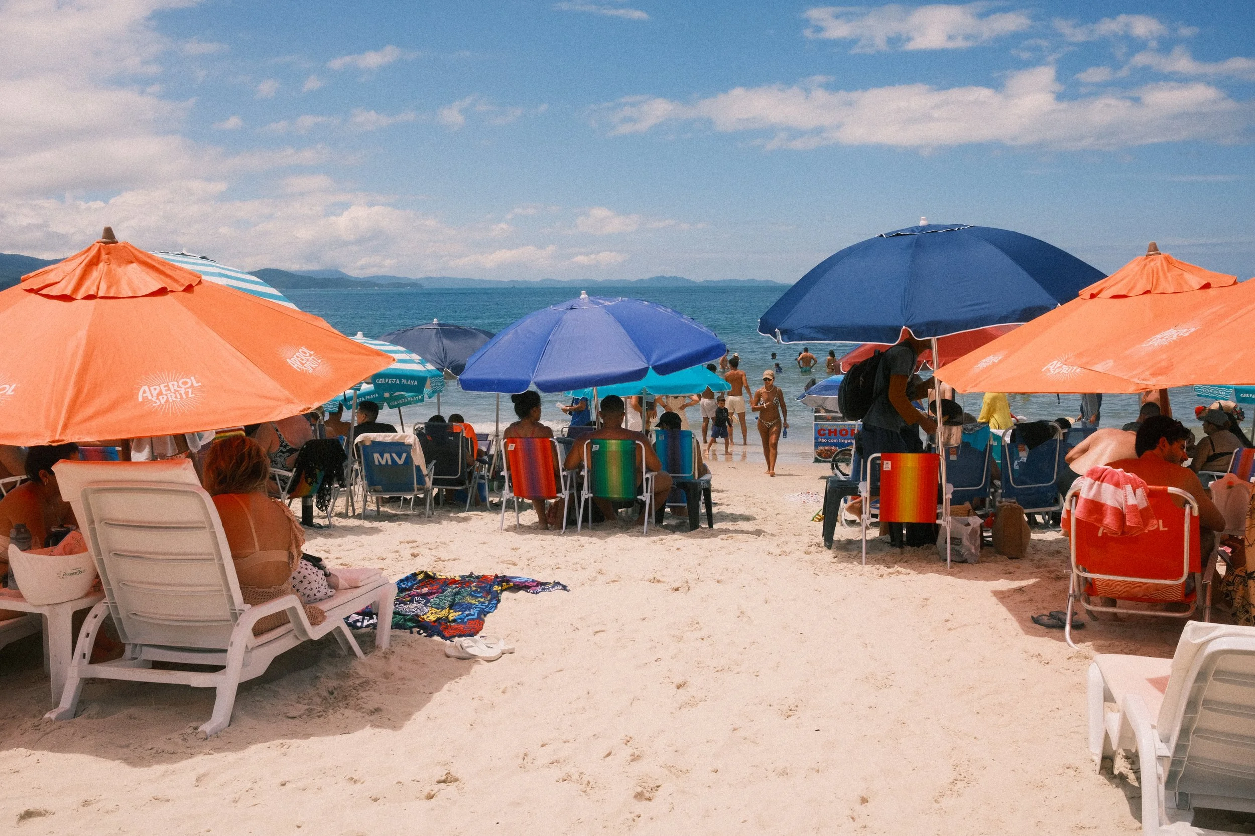 People relaxing on a crowded beach under colorful umbrellas with the ocean and blue sky in the background.