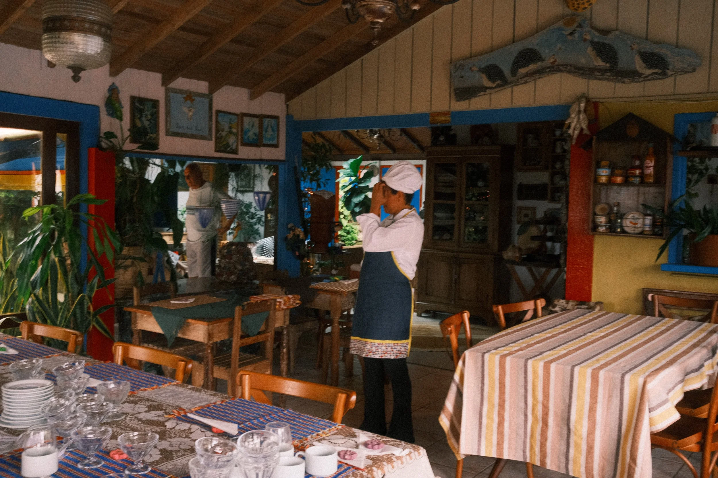 A woman dressed as a chef standing in a rustic restaurant with wooden chairs, tables, and colorful decorations