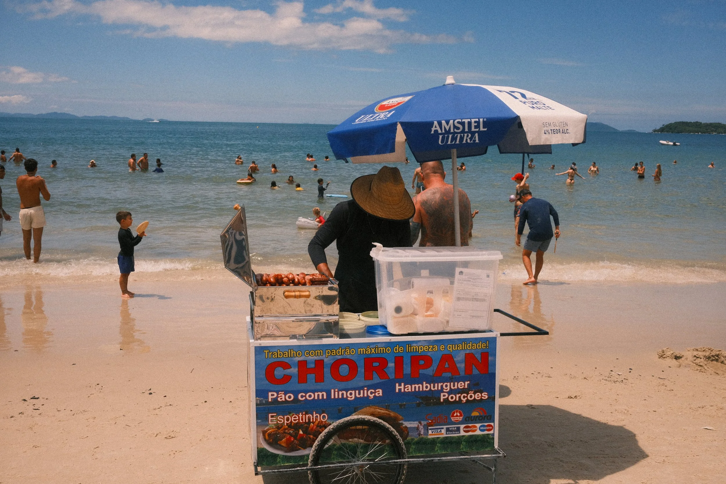 Beach scene with people swimming and playing in the water, a hot dog cart on the sandy shore, and an umbrella providing shade, with a scenic ocean view in the background.