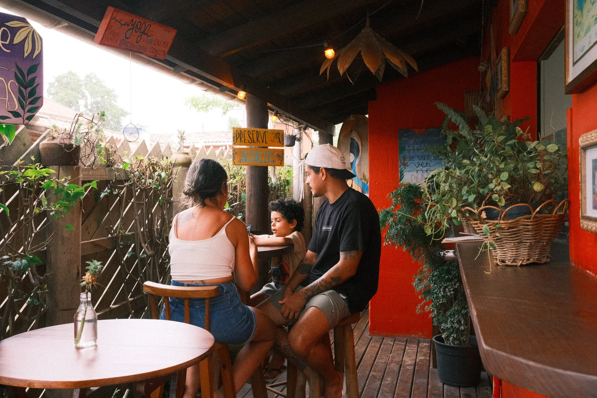 A family sitting at a table in a cozy, decorated outdoor cafe, with plants and colorful wall art surrounding them.